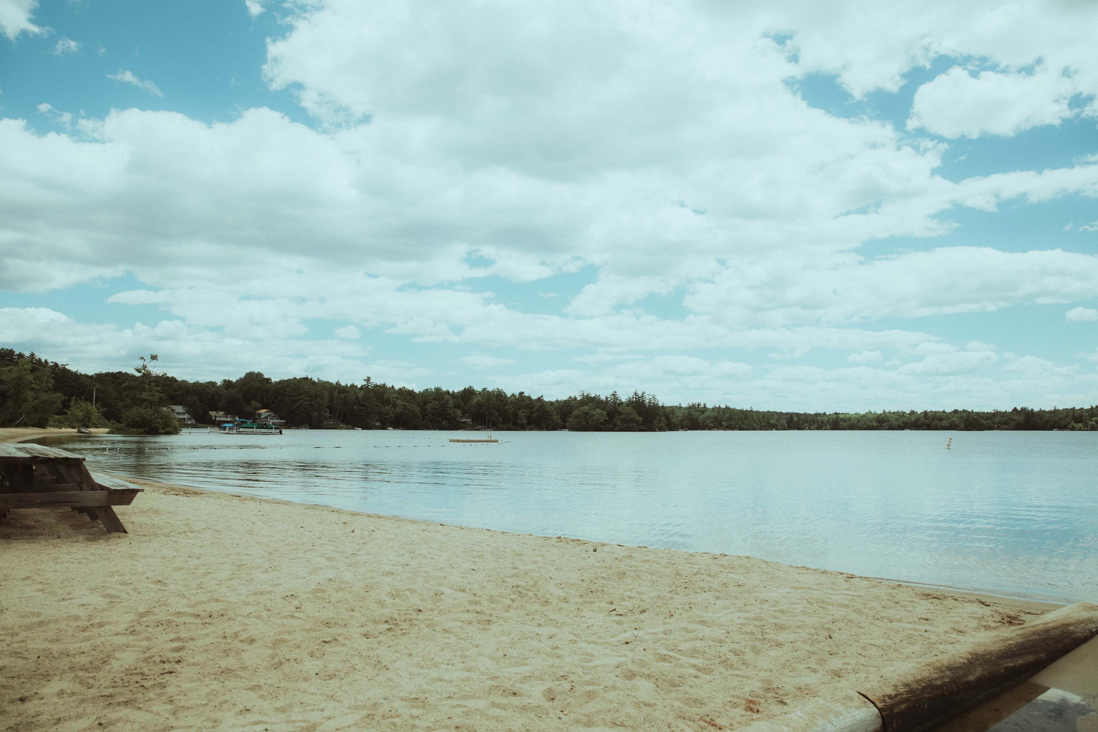 A sandy beach beside a calm lake with scattered clouds in a blue sky and trees lining the shoreline.