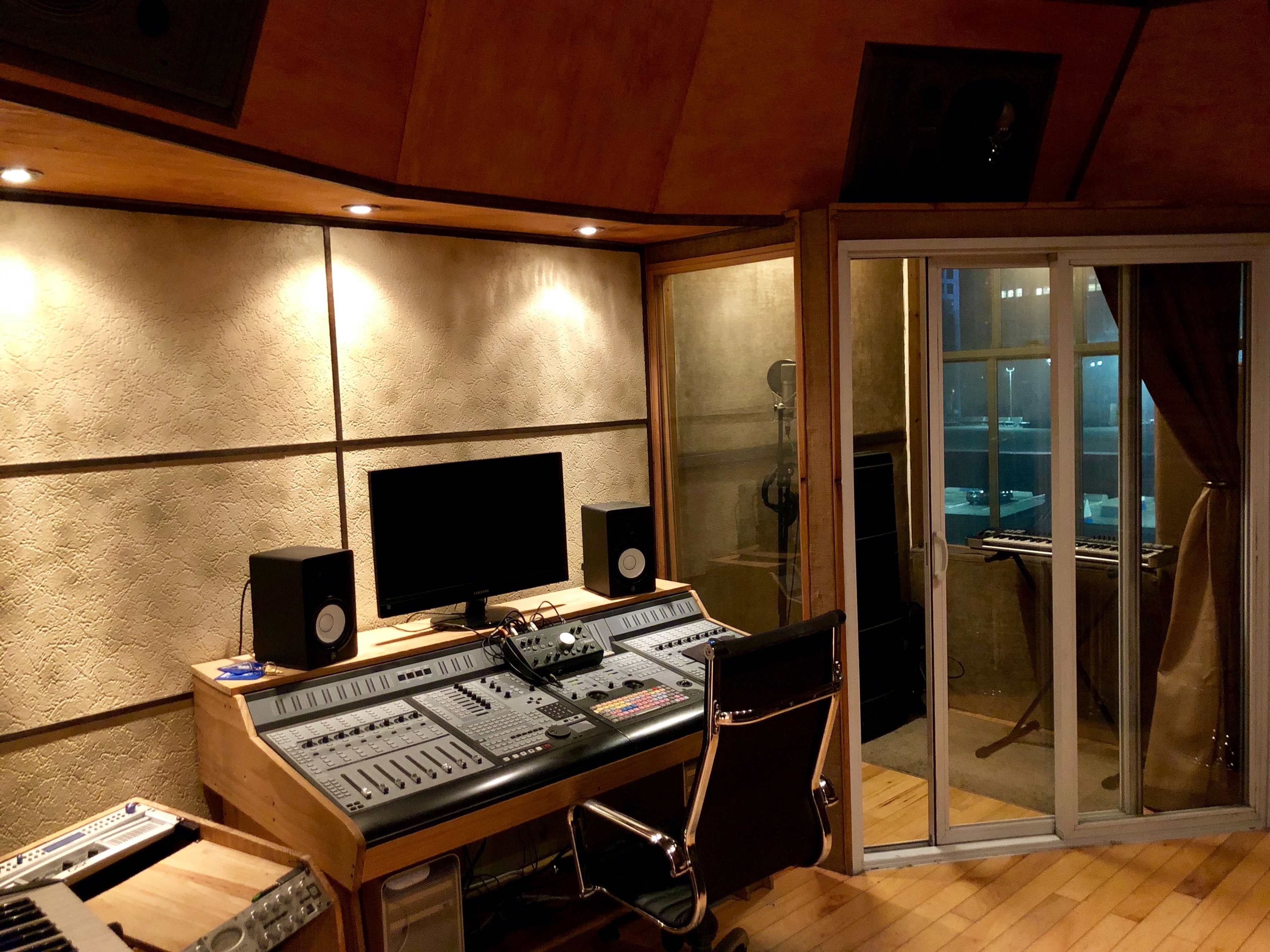 The image shows a music recording studio featuring a control desk with a computer monitor, speakers, and a glass-walled vocal booth.