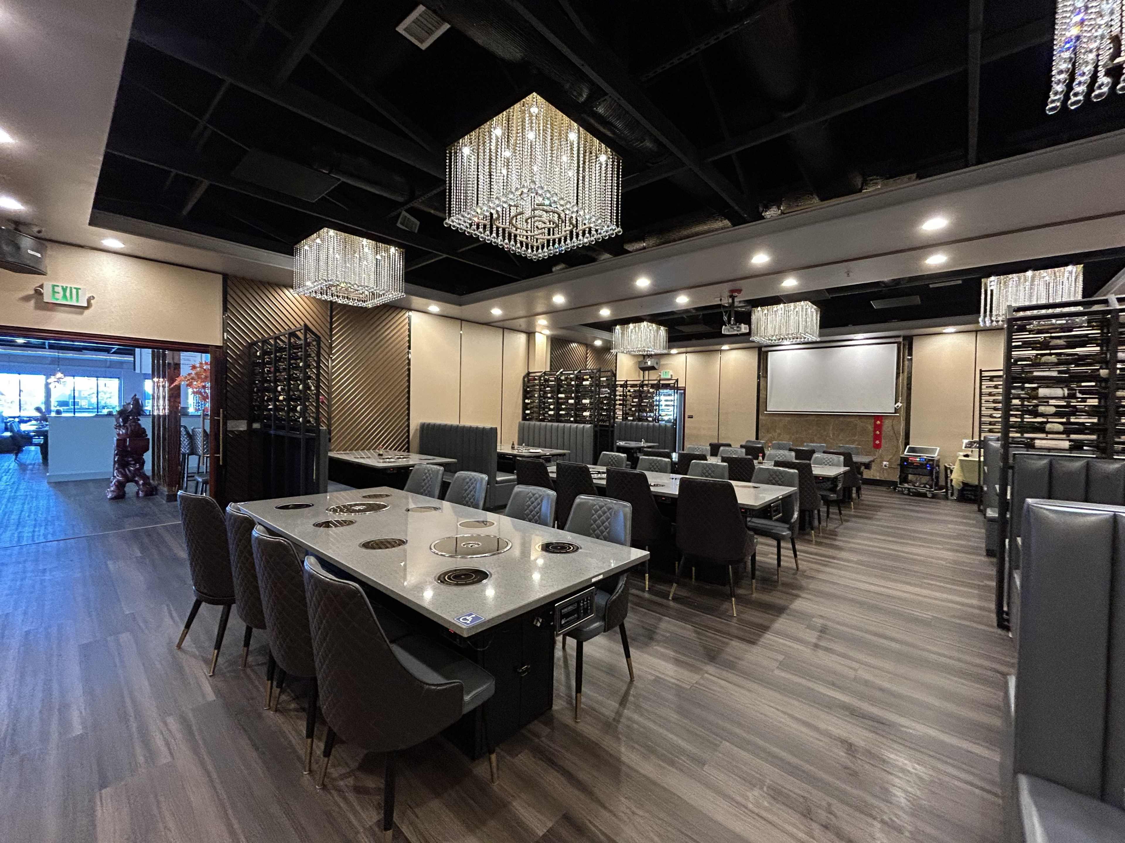 A modern restaurant interior featuring tables with built-in grilling stations, elegant chandeliers, and a wine rack in the background.