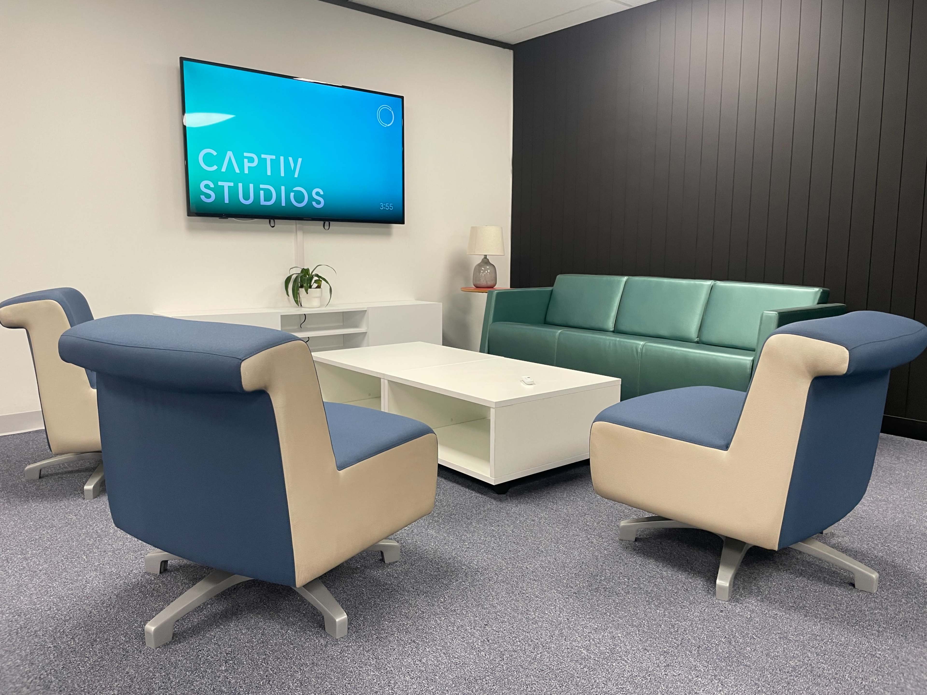 A modern seating area with a teal sofa, light-colored armchairs, a white coffee table, and a wall-mounted television displaying "CAPTIV STUDIOS."