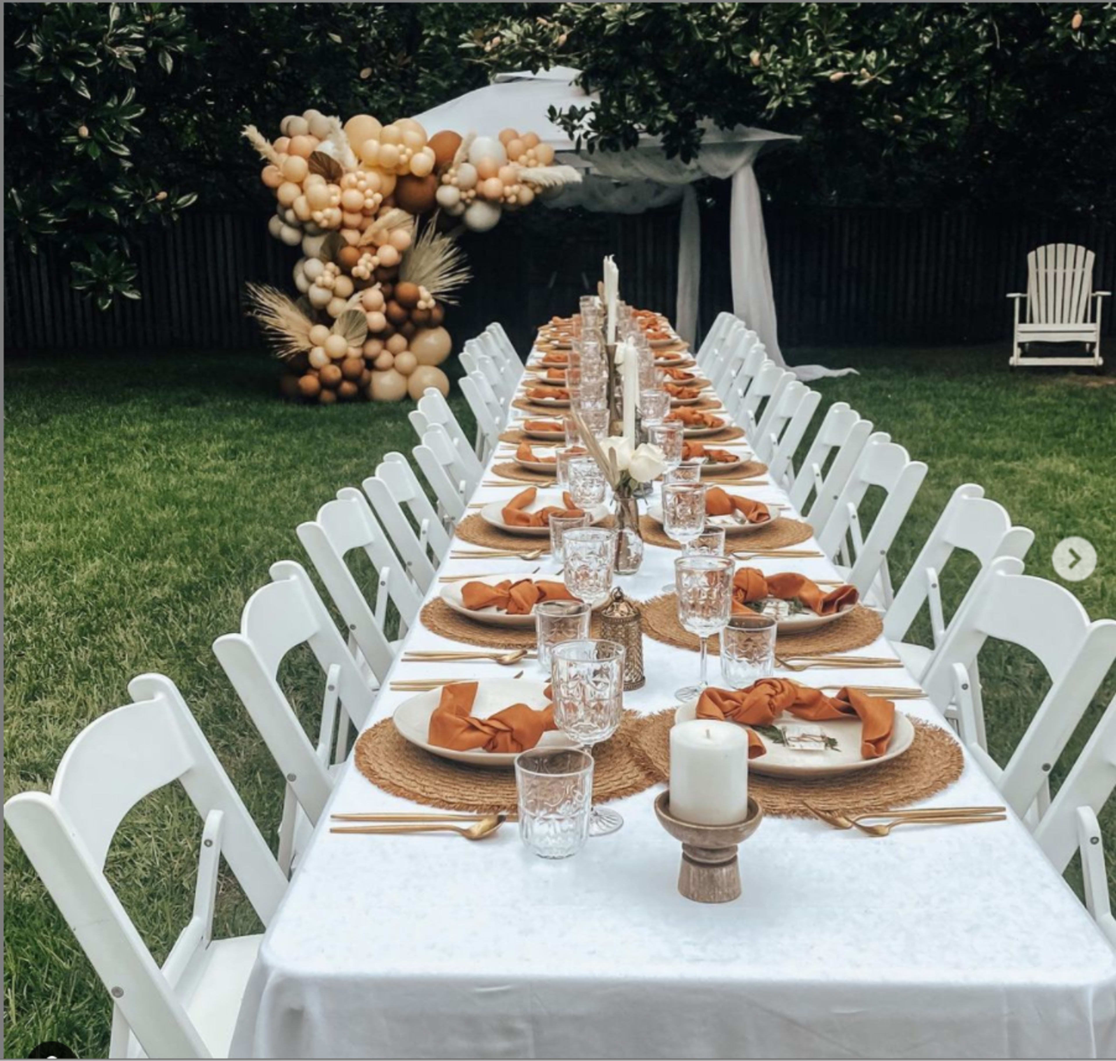 A long, elegantly set outdoor dining table is arranged on a green lawn, flanked by white chairs and decorated with neutral-toned tableware and a backdrop of a balloon arch.