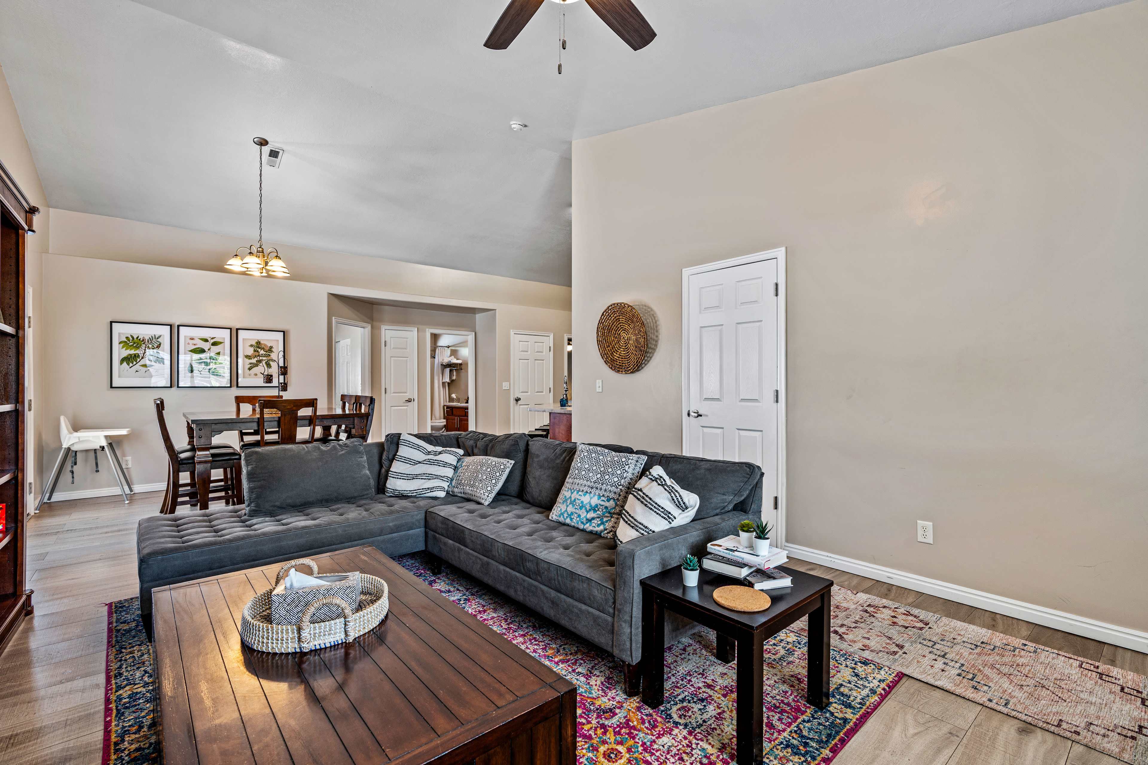 The living area features a dark sectional sofa, a wooden coffee table, and a dining area visible in the background.
