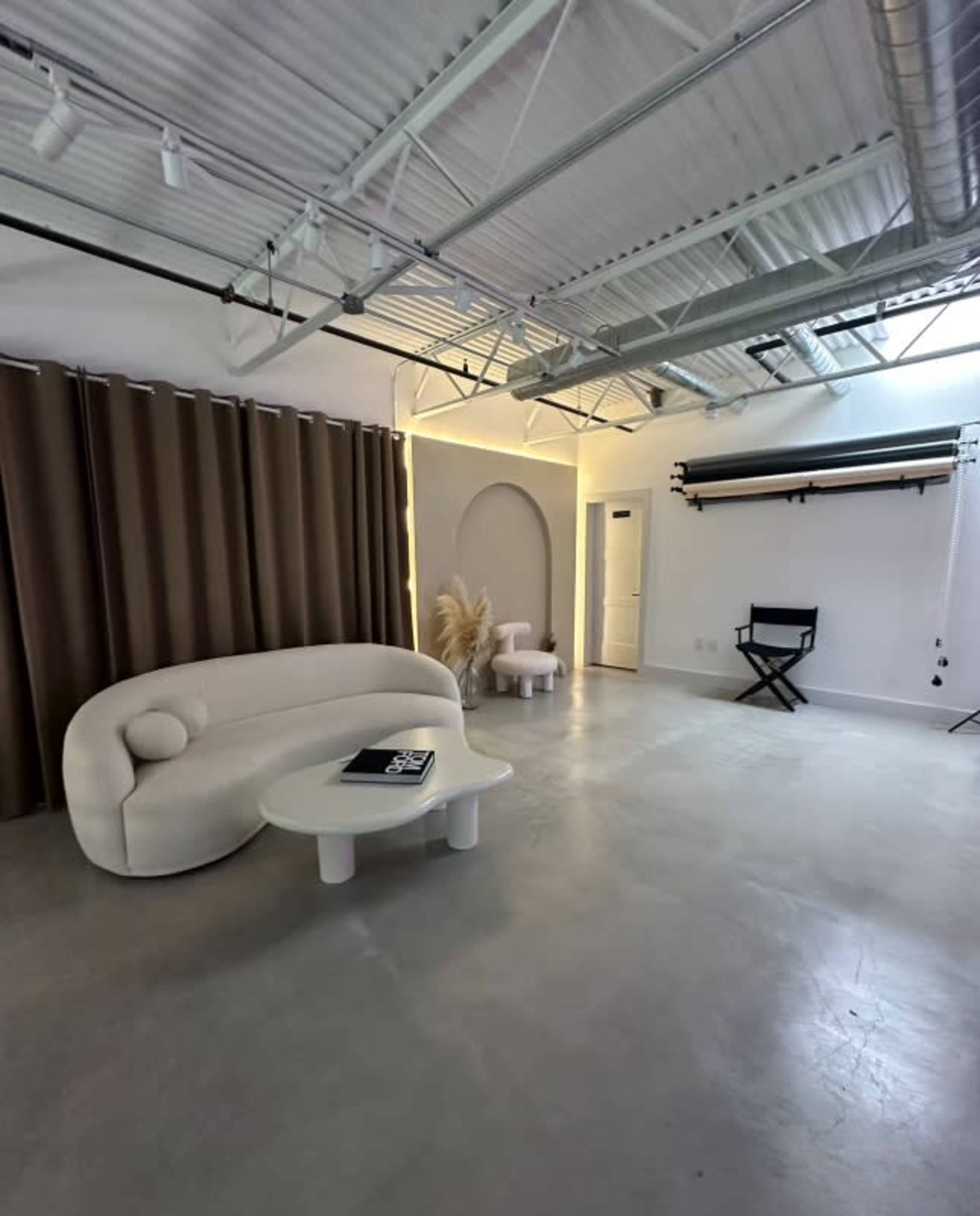 A spacious, minimalist studio with a curved white sofa, a round table, and large curtains in muted tones.