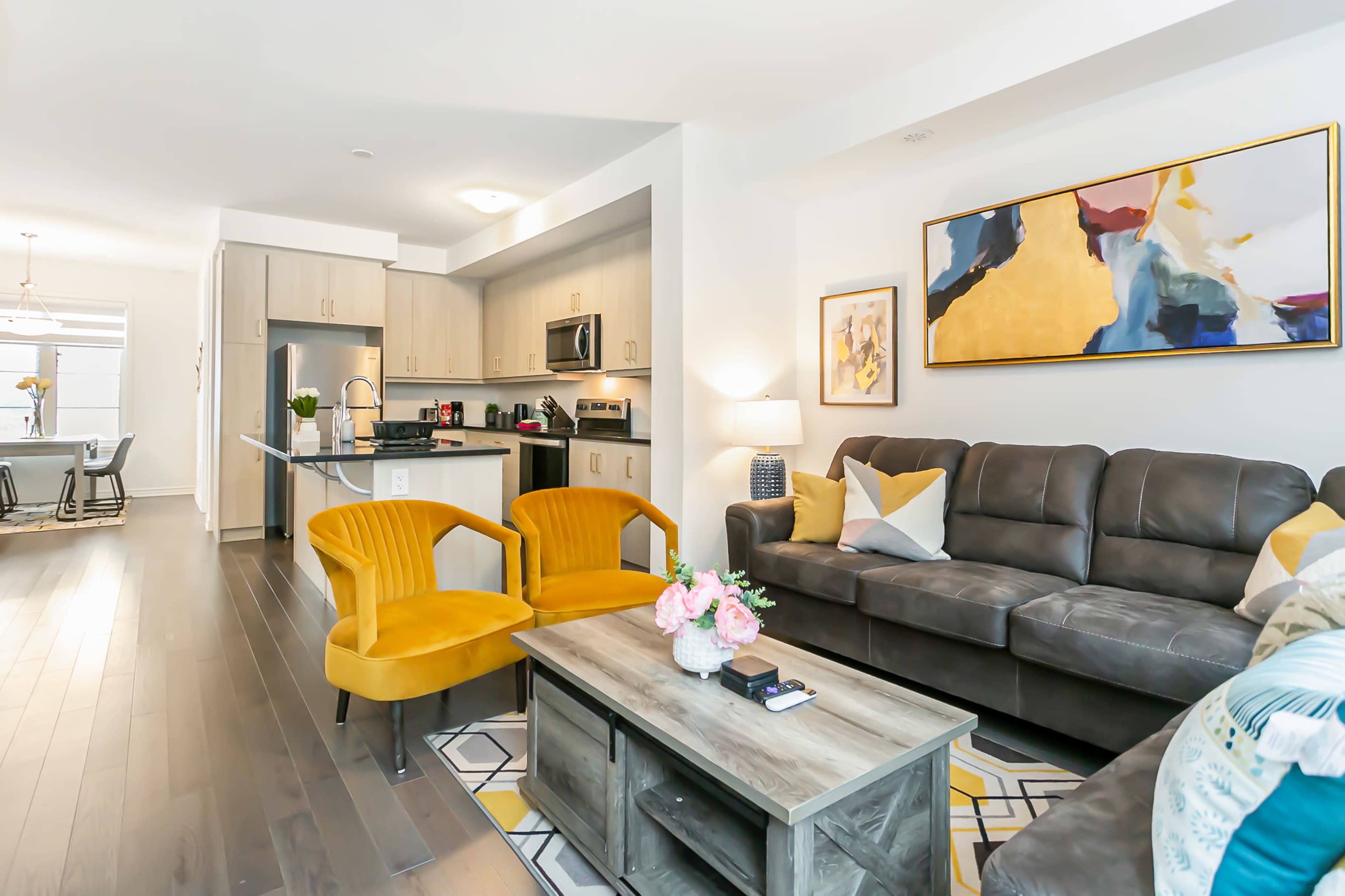 A modern living room with a gray sofa, yellow accent chairs, a wooden coffee table, and an adjoining kitchen area with sleek cabinets and stainless steel appliances.