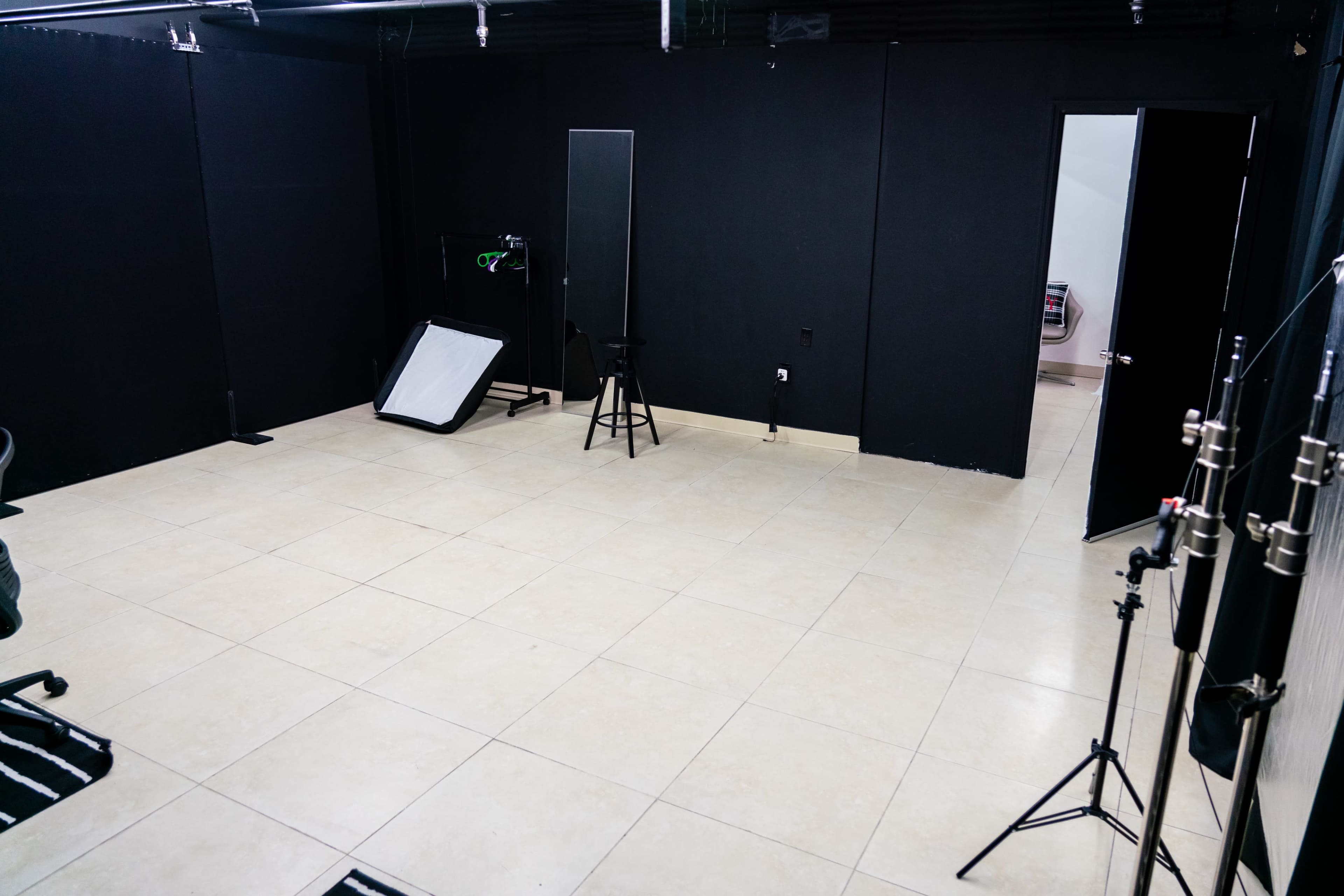 The image shows an empty studio space with black walls, a large mirror, a stool, and lighting equipment.