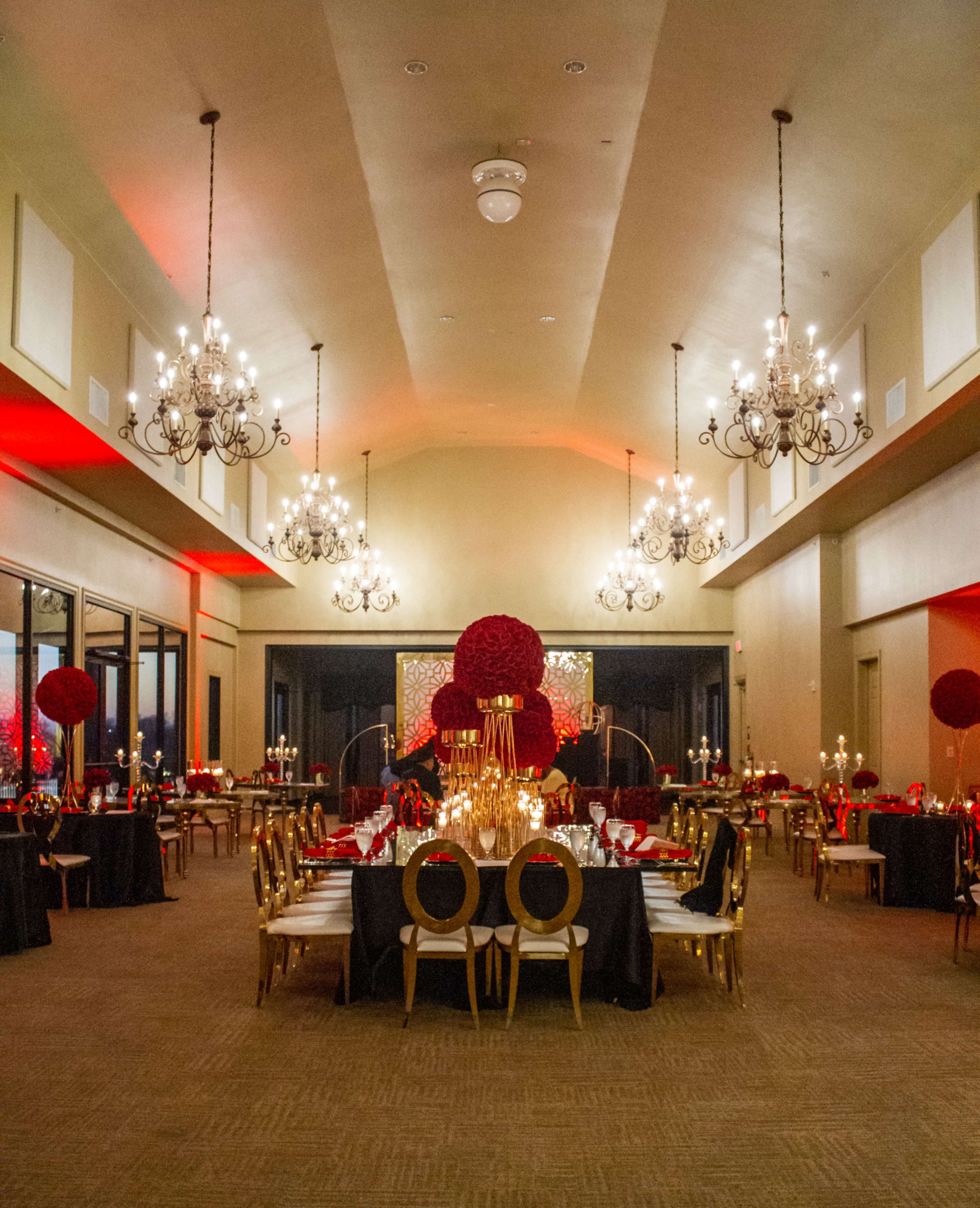 The image shows a large banquet hall decorated for an event, featuring tables set with red and black tablecloths, tall floral arrangements, and elegant chandeliers.