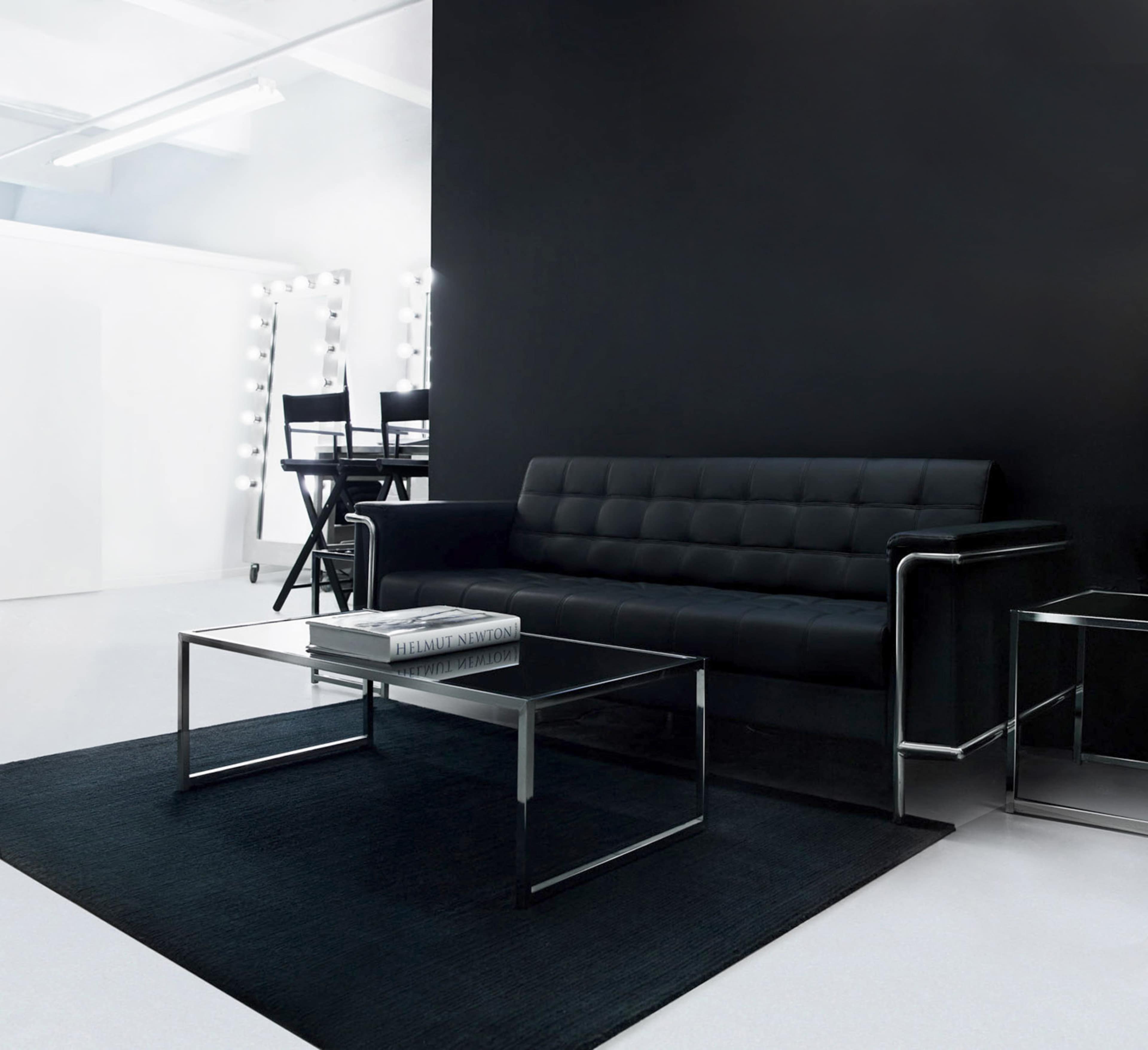 A modern black sofa positioned in front of a black wall, accompanied by a glass coffee table and a dressing table with lights in the background.