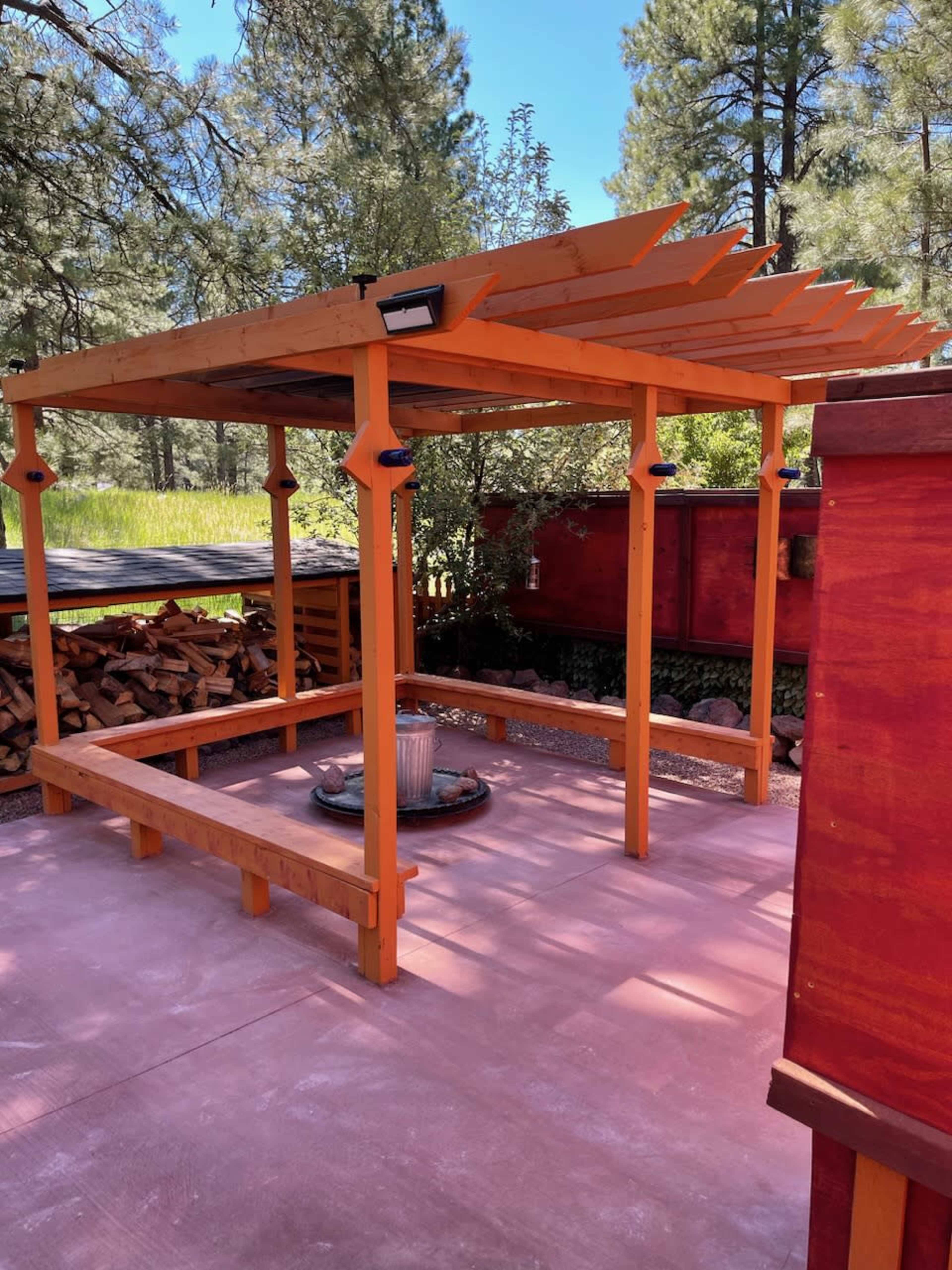 A wooden pergola with an open roof, situated on a concrete platform surrounded by stacked firewood and trees.