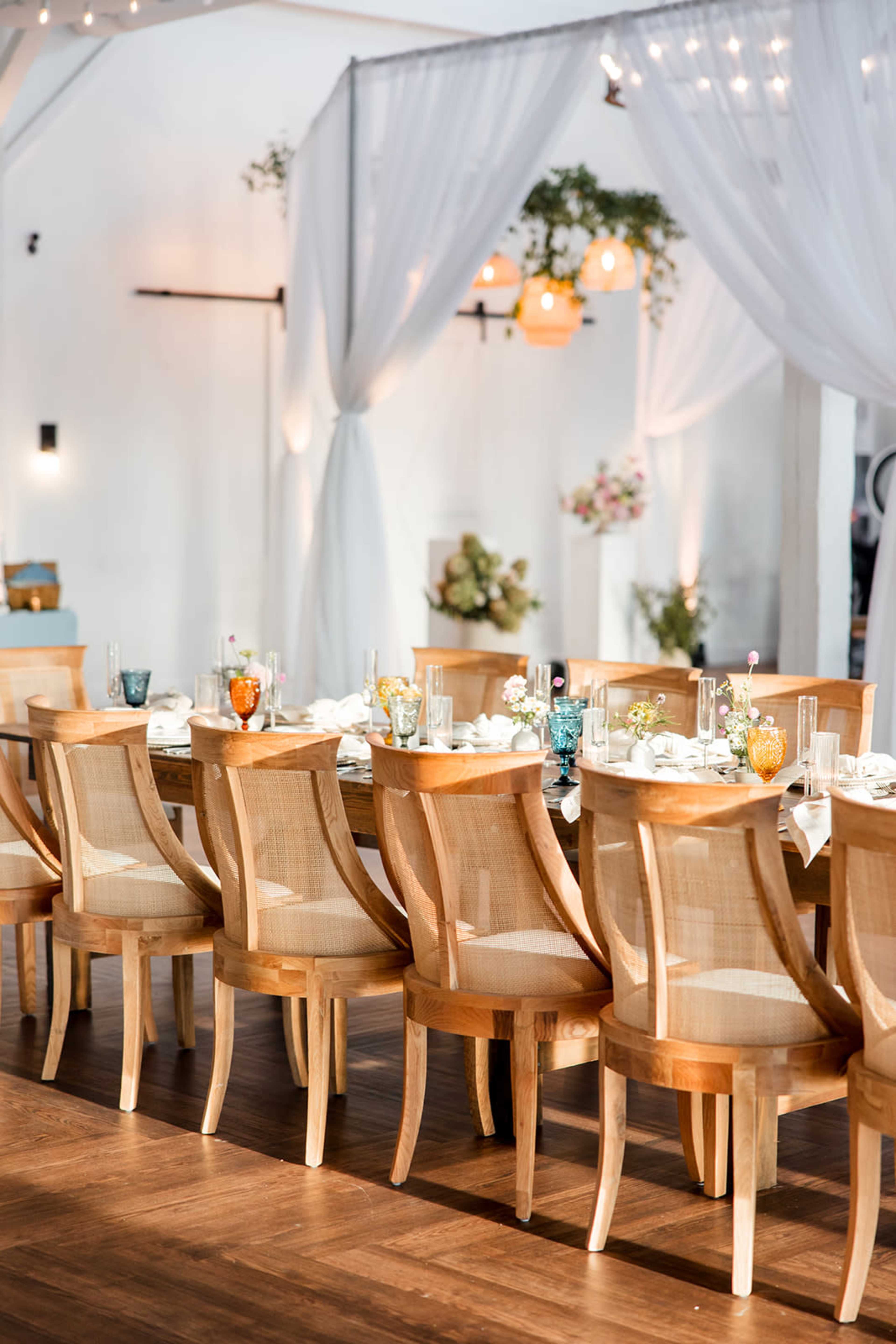 A long wooden dining table is set with white tableware and surrounded by multiple wooden chairs in a decorated event space with soft lighting and draped fabric.