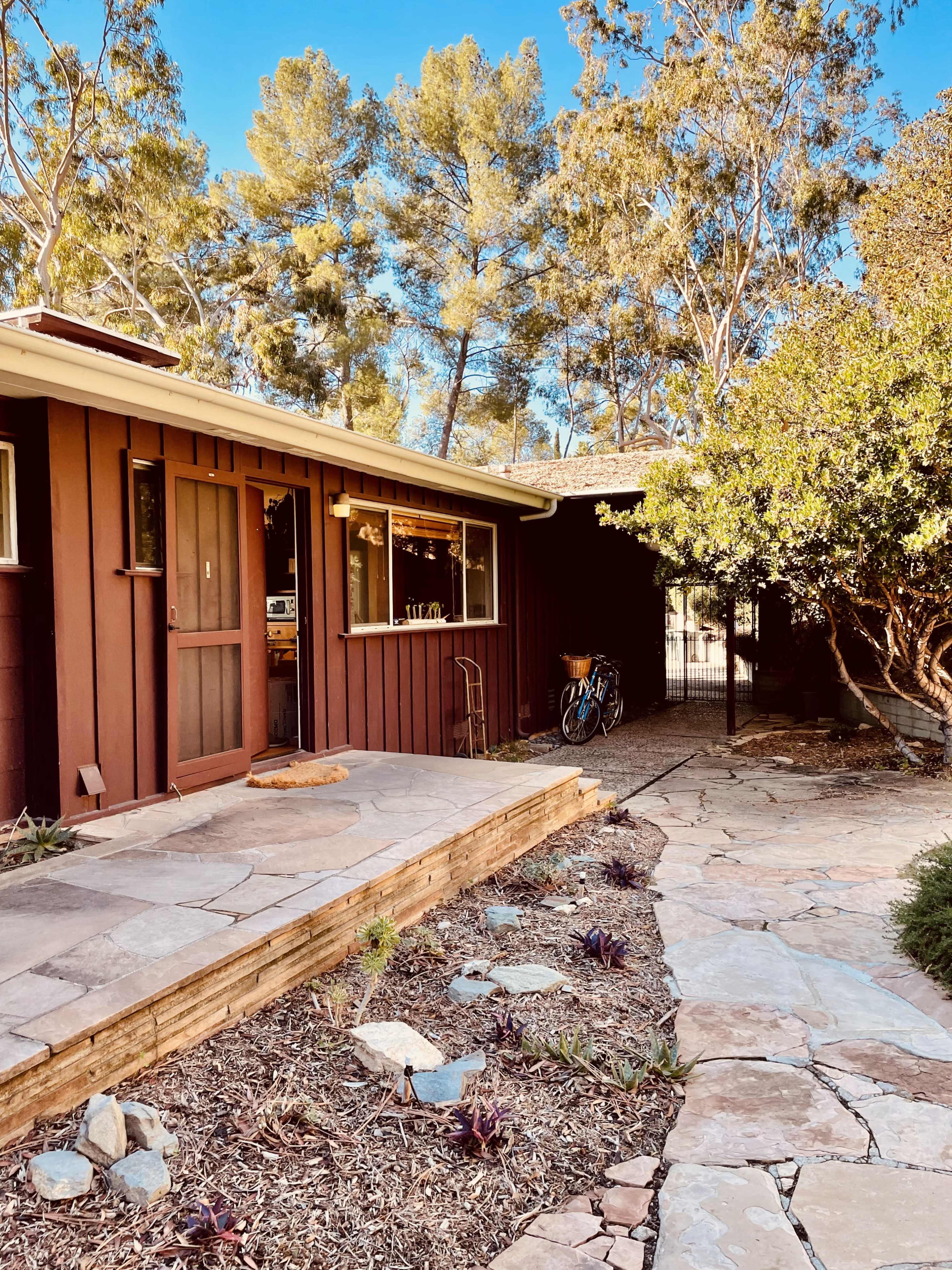 The image shows a brown wooden house with a stone pathway leading up to it, surrounded by trees and shrubs.