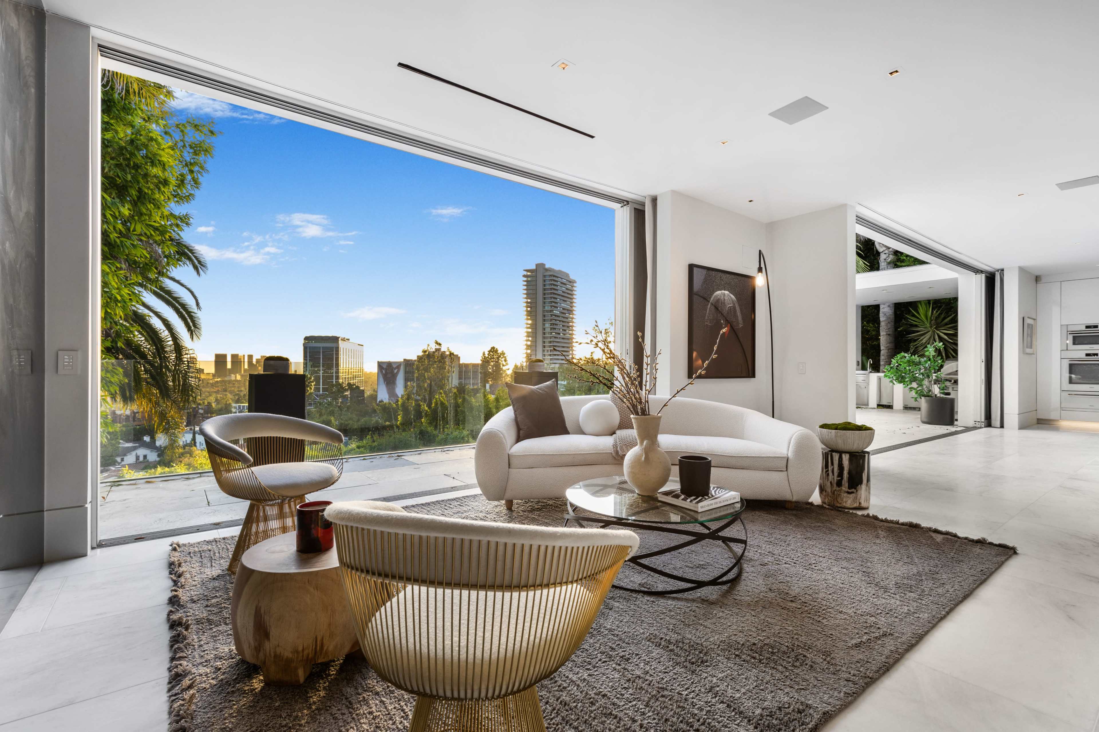 The image shows a modern living room with a white sofa and a round coffee table, featuring large glass doors that open to a view of a city skyline and greenery.