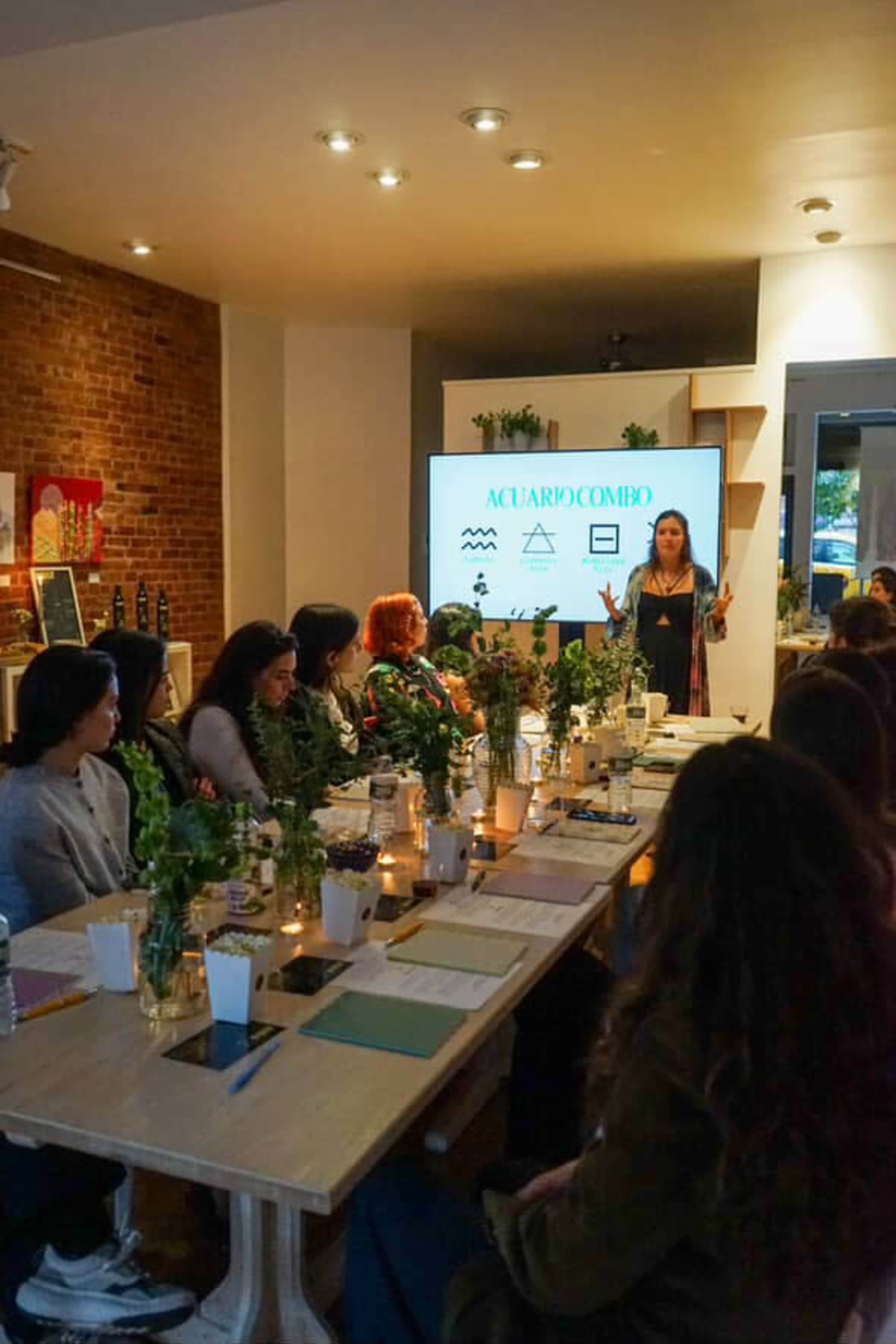A woman presents in front of a group seated at a long table decorated with green plants and candles in a well-lit room.