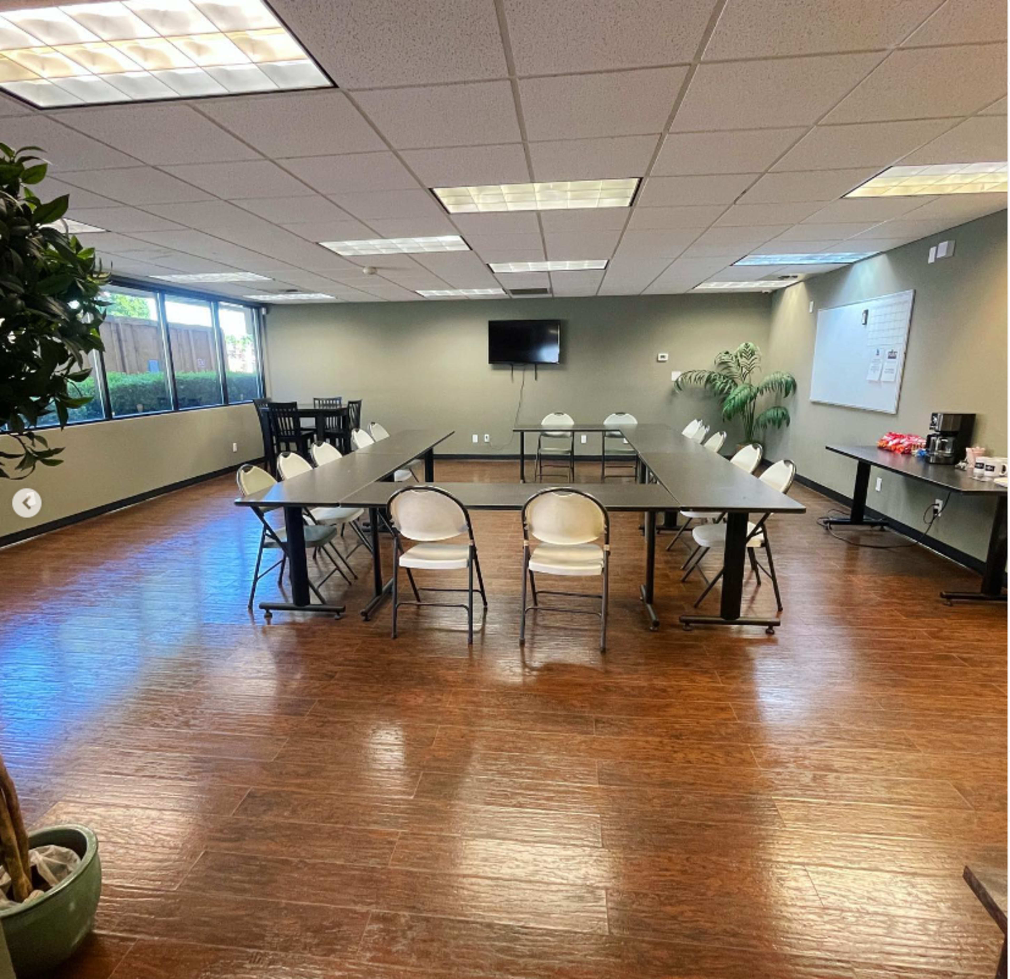 A large, minimally furnished meeting room features a long table surrounded by chairs, with windows allowing natural light and a television mounted on the wall.