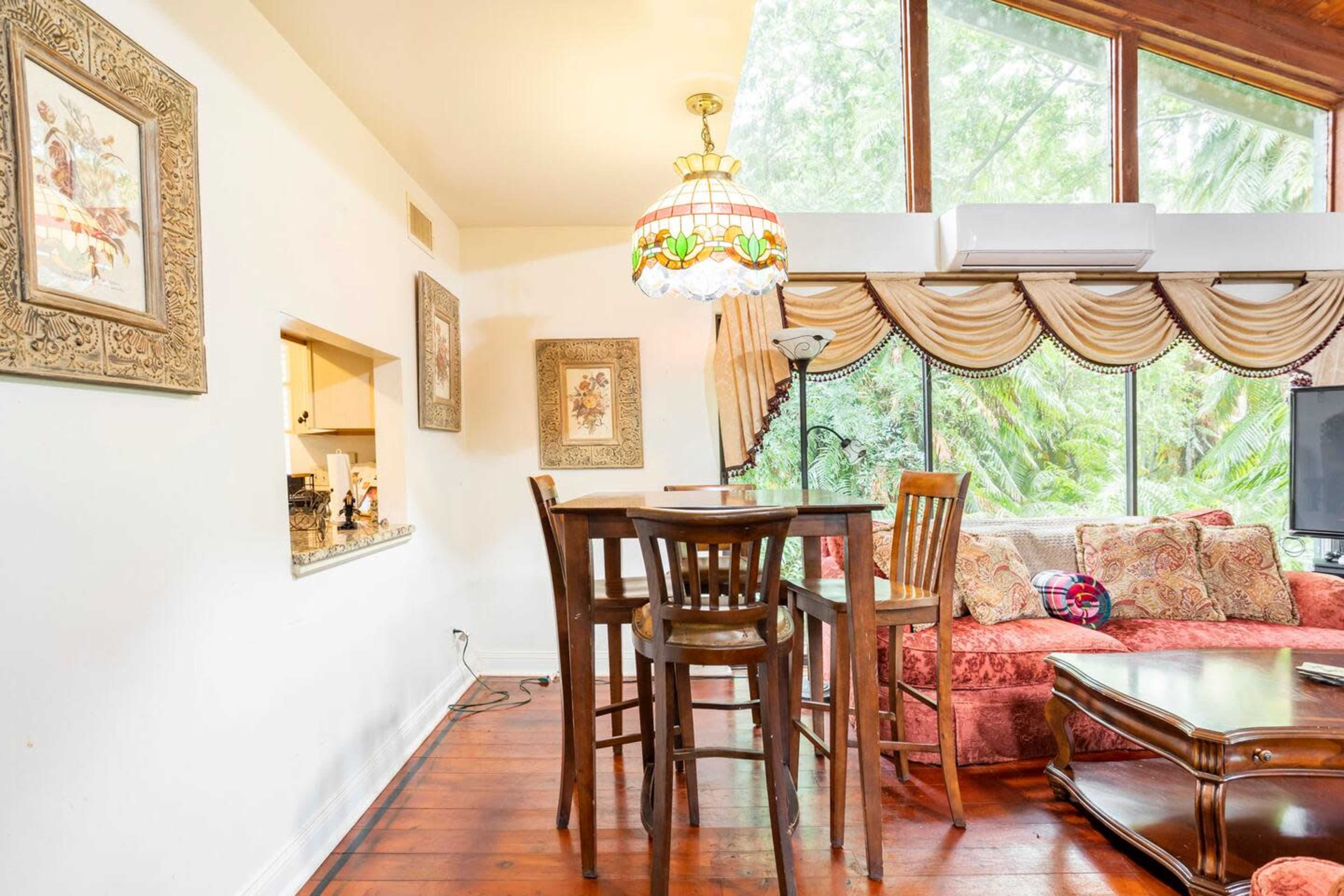 The image depicts a dining area with a wooden table and chairs adjacent to a living space featuring a sofa and large windows overlooking greenery.