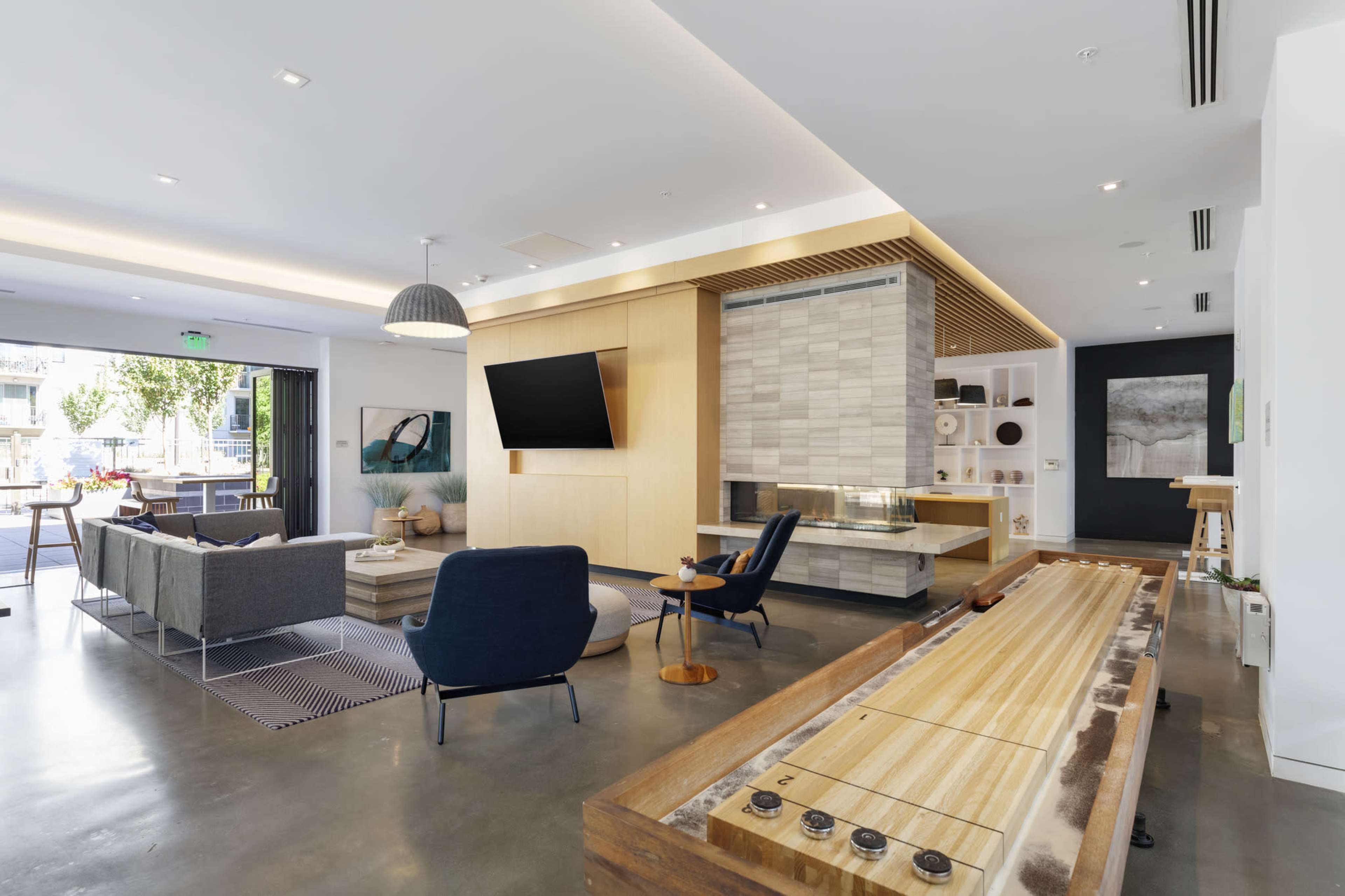 A modern living room featuring a shuffleboard table, a seating area with chairs and sofas, and a large flat-screen TV mounted on a wooden wall.