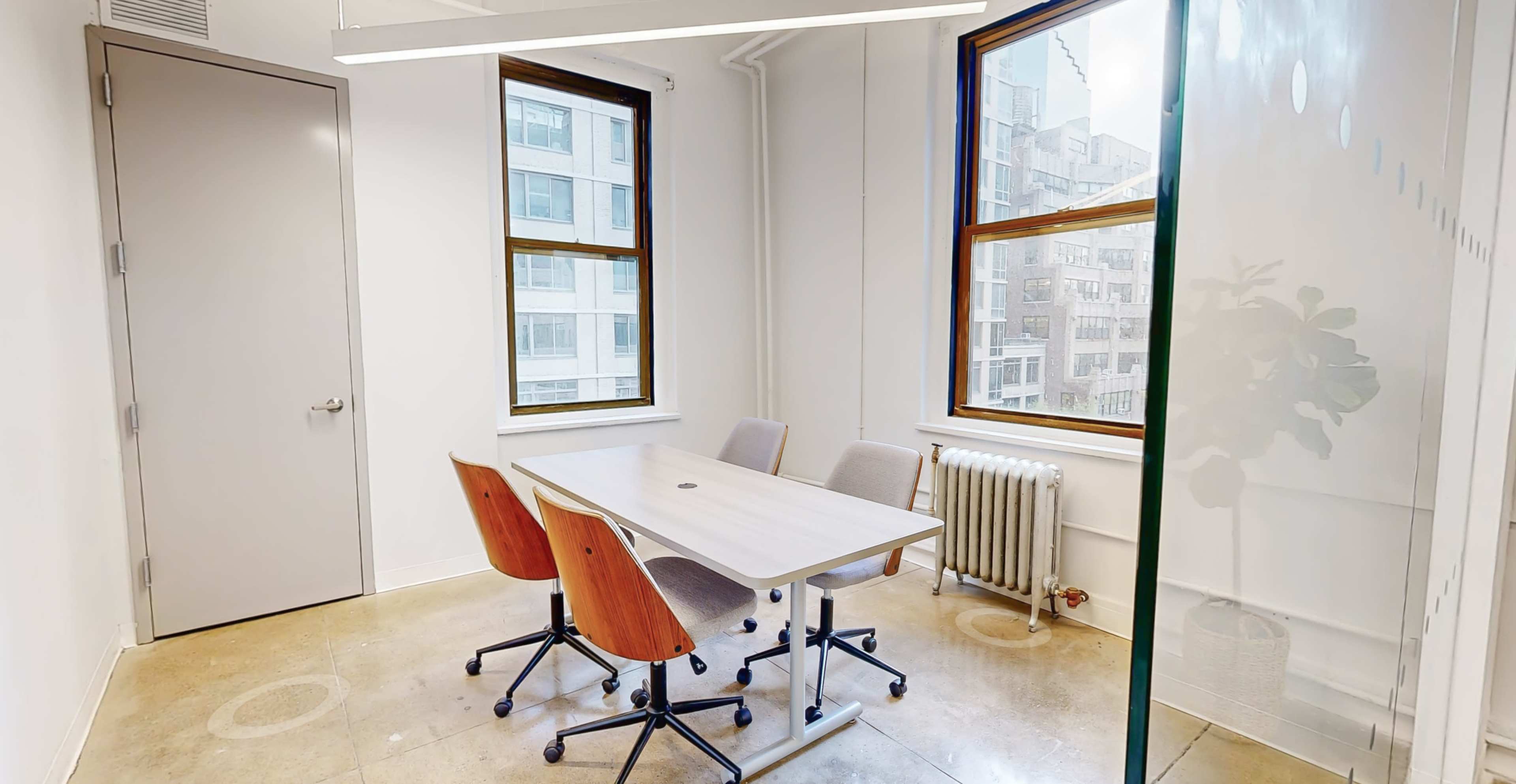 A small meeting room with a table and four chairs, featuring large windows that let in natural light and a radiator along one wall.