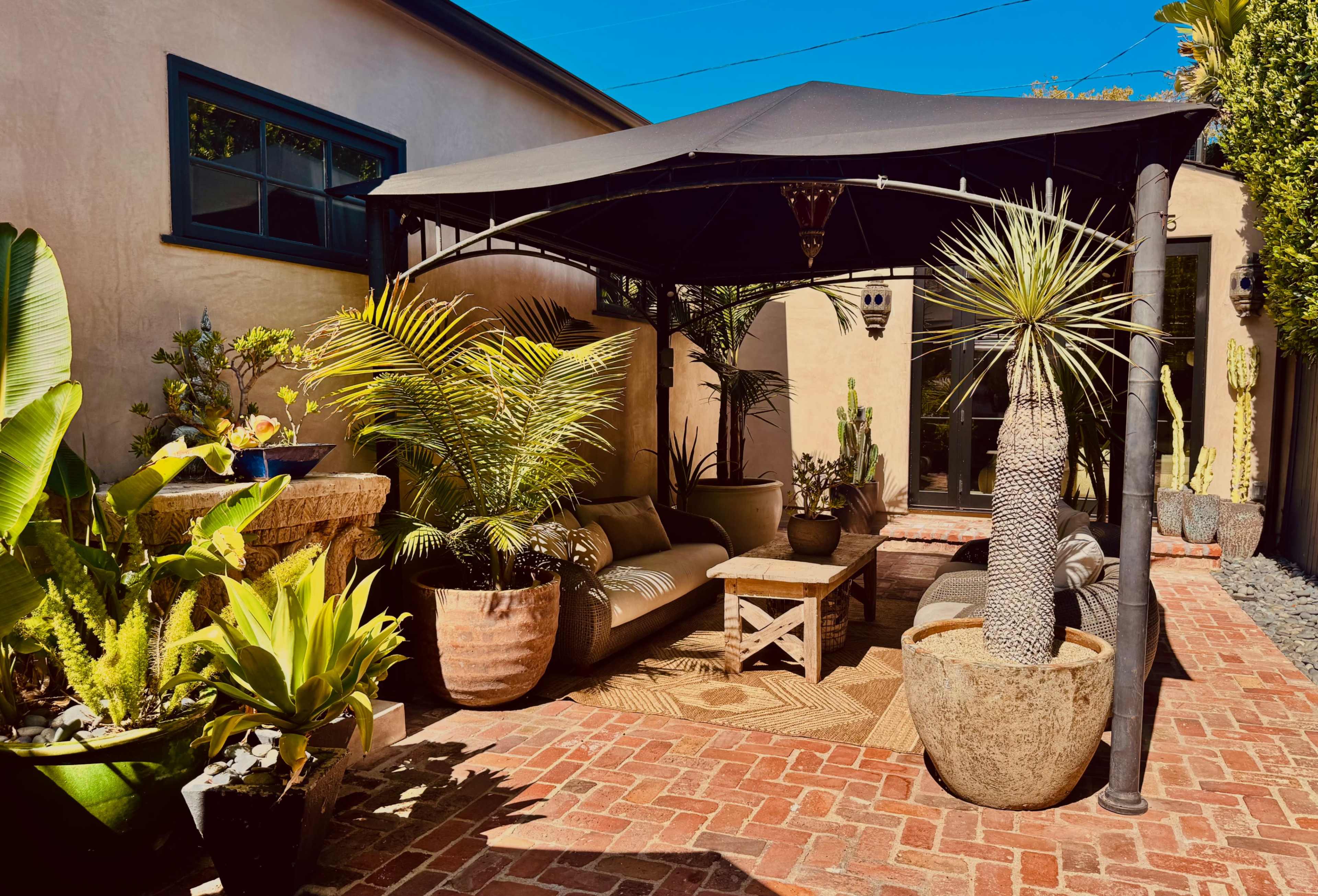 A shaded outdoor seating area features a black canopy, comfortable furniture, and various potted plants, surrounded by brick flooring.