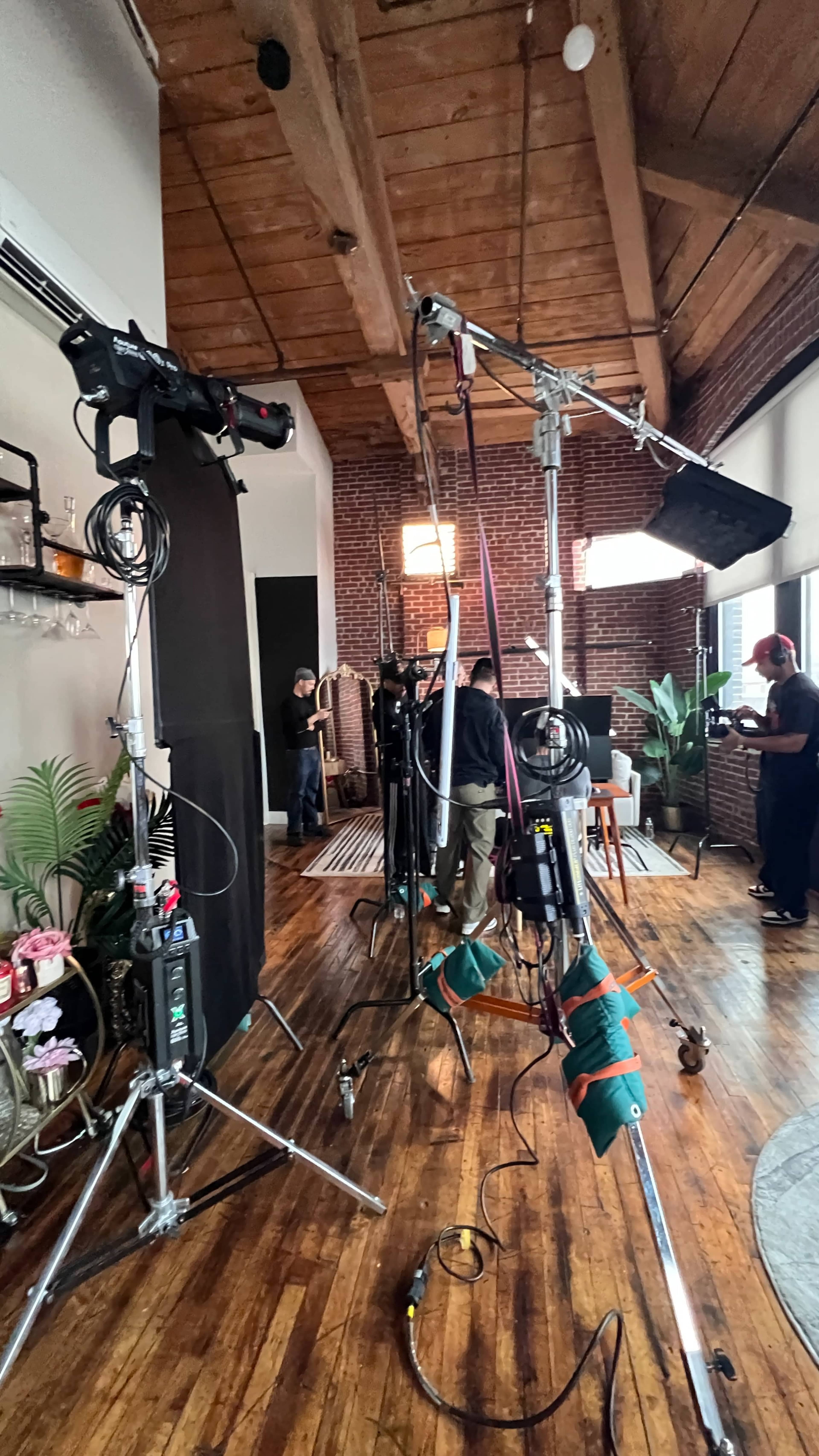 A film set is arranged with various cameras, lighting equipment, and crew members working in a room with wooden flooring and brick walls.