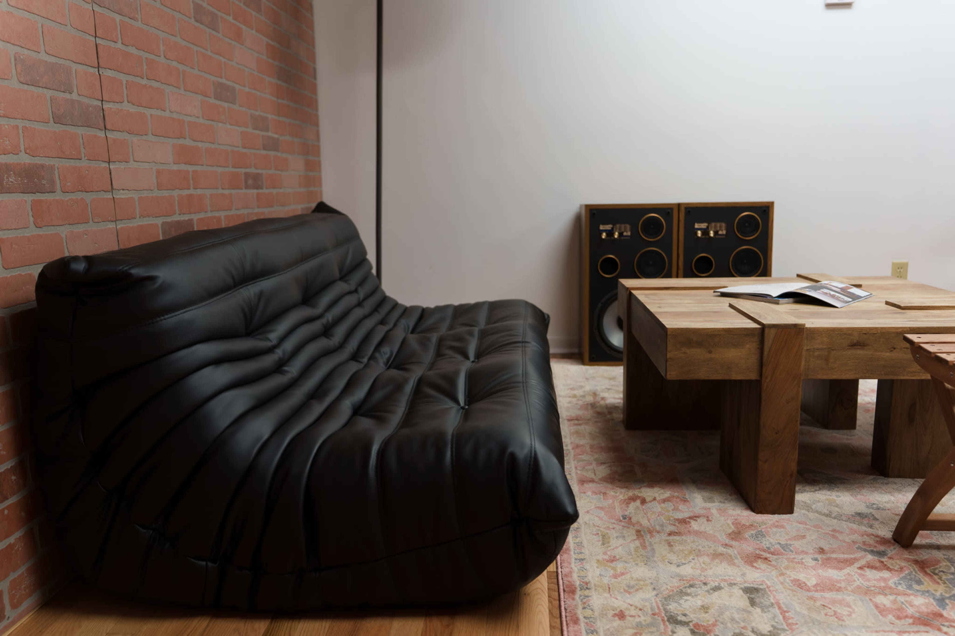 The image shows a black, tufted sofa positioned against a brick wall, next to a wooden coffee table and a sound system with large speakers.