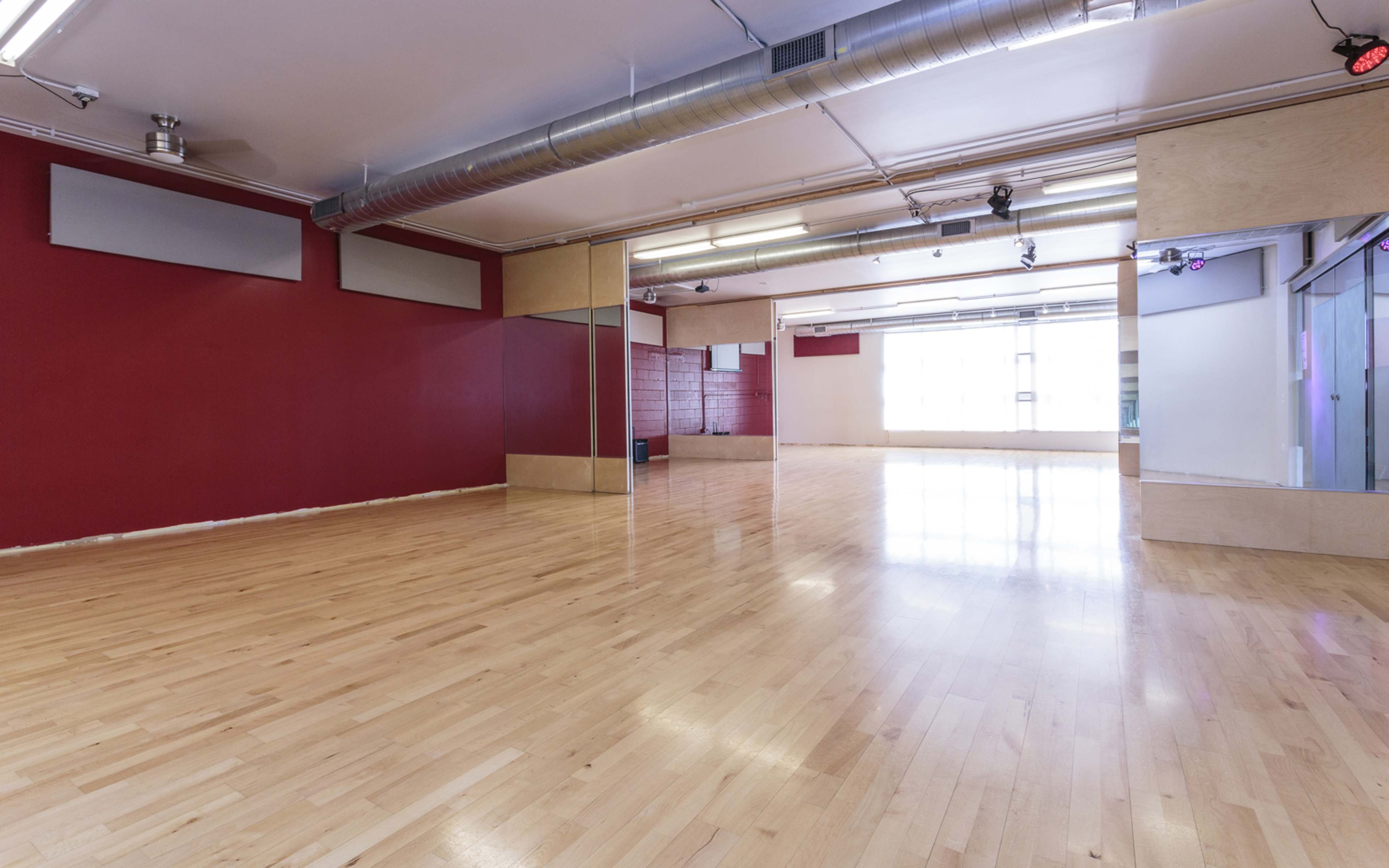 A spacious dance studio with hardwood floors and red and light-colored walls, featuring large windows and overhead lighting.