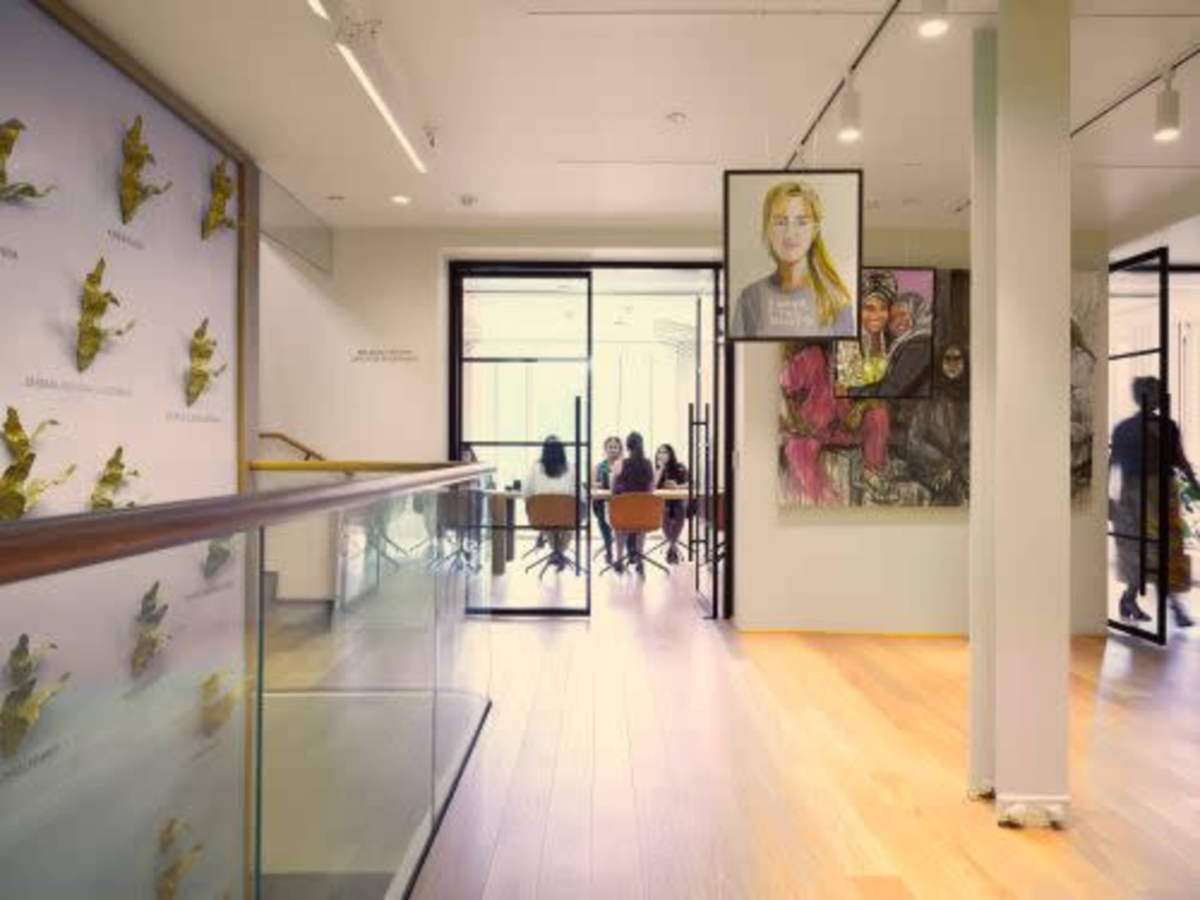 The image shows a modern interior space with a glass railing, featuring contemporary artwork on the walls and a group of people seated at a table in a meeting area.