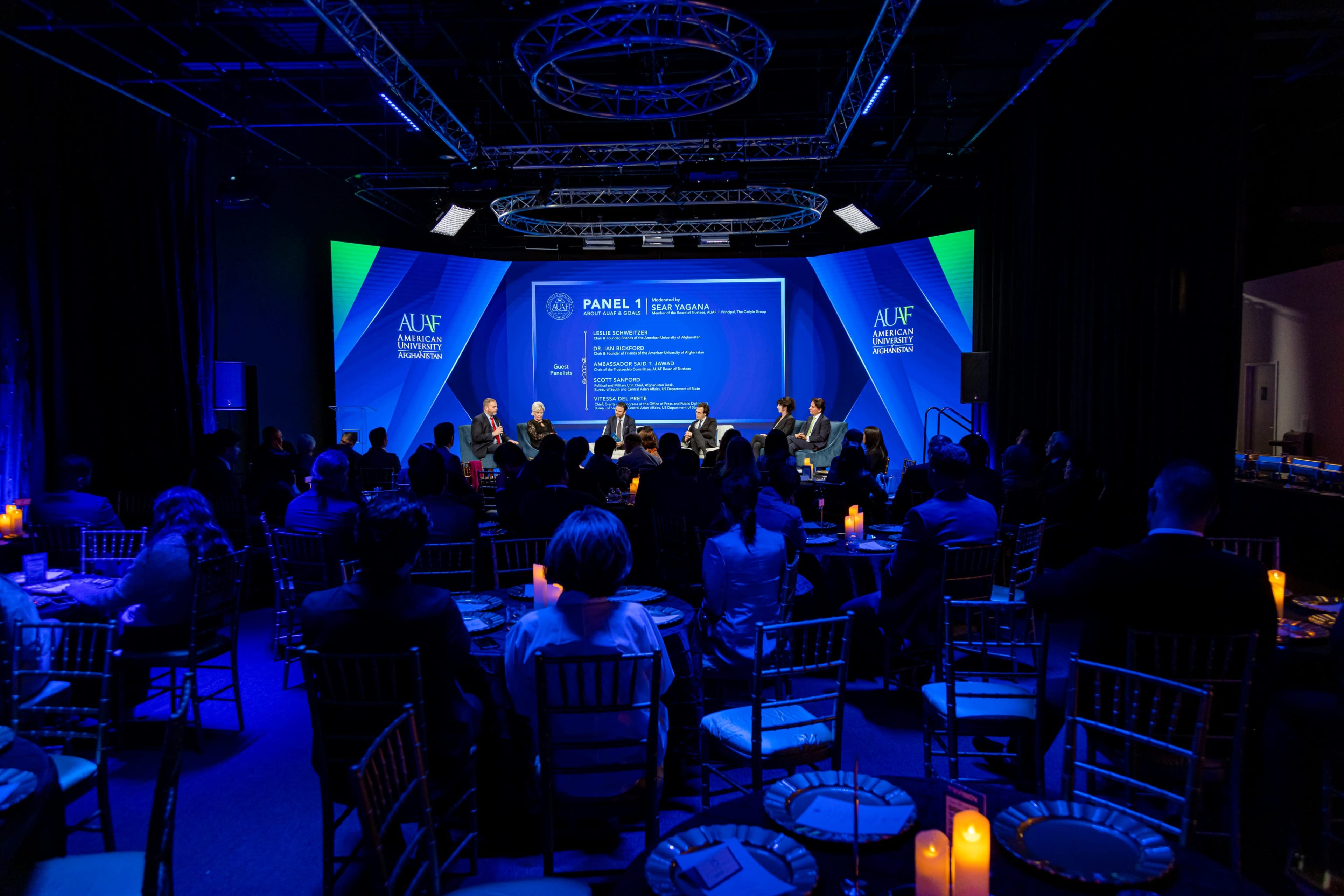 A panel discussion is taking place on stage, with speakers seated in front of an audience, illuminated by blue lighting in a conference setting.