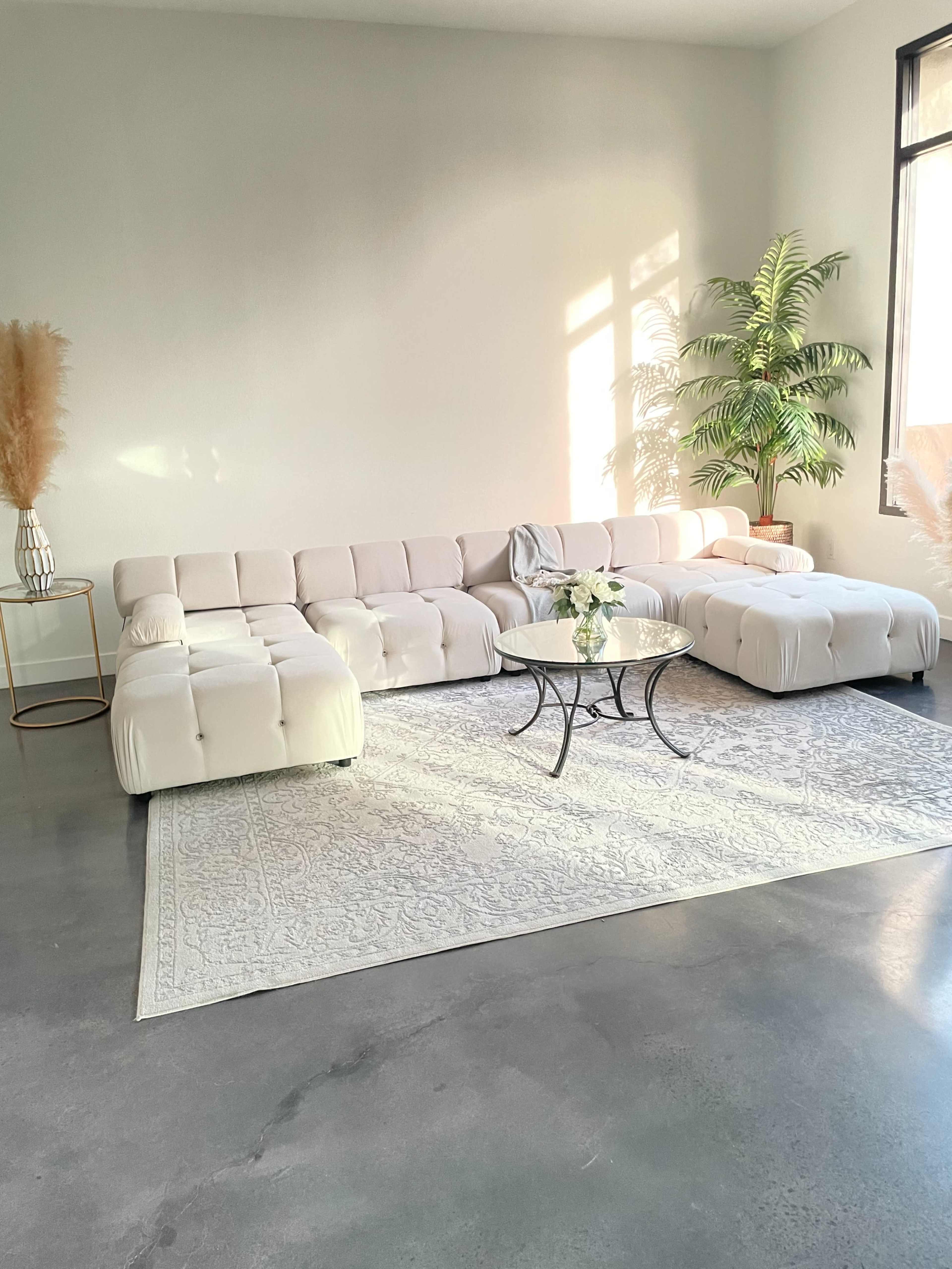 The image shows a modern living room with a large white sectional sofa, a glass coffee table on a light gray rug, and a potted plant by the window.