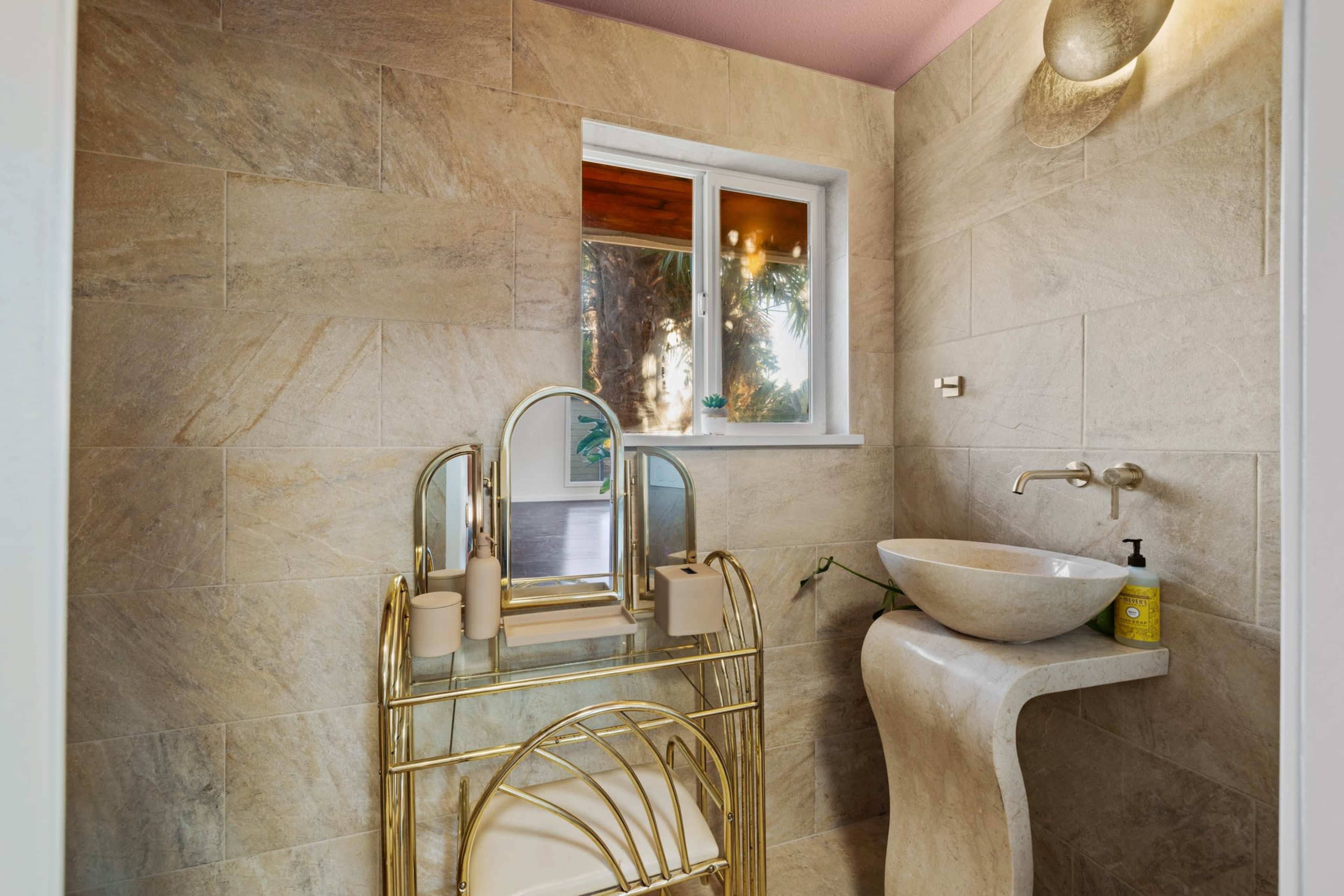 A bathroom with stone tile walls, a round basin on a curved countertop, and a decorative gold-framed mirror on a gold cart.