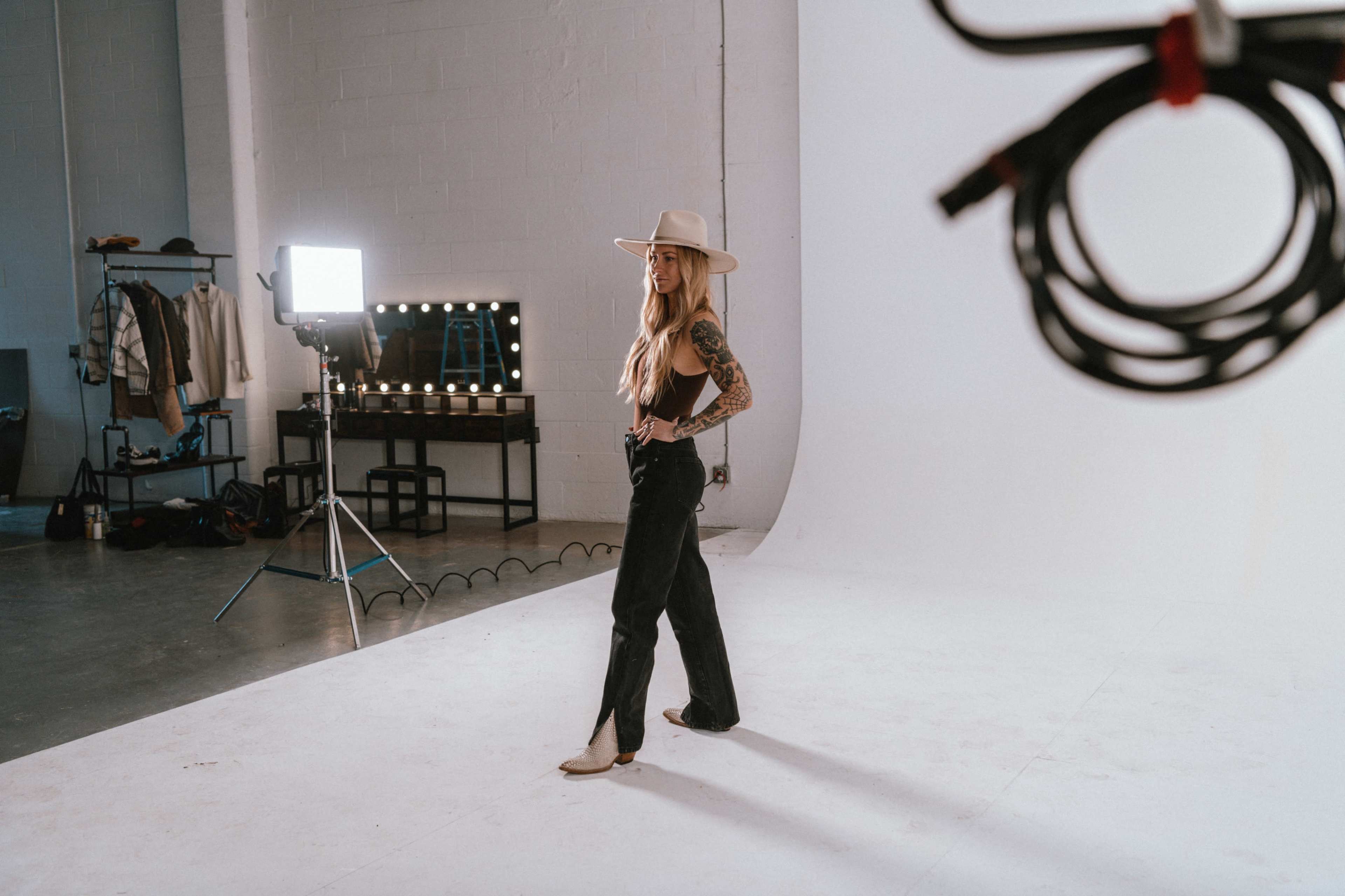 A woman with long hair and tattoos poses in a photography studio with a white backdrop and soft lighting equipment.