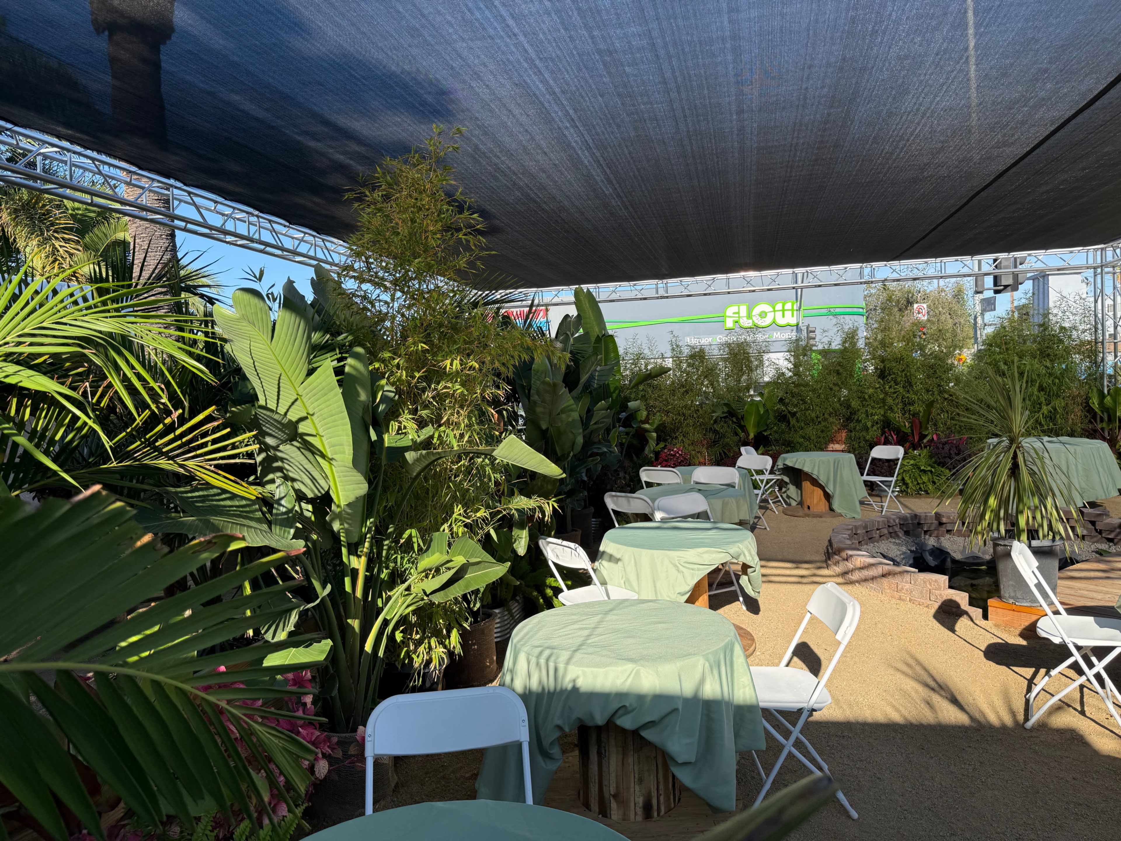 The image shows a shaded outdoor area filled with tables covered in green cloth, surrounded by various tropical plants.