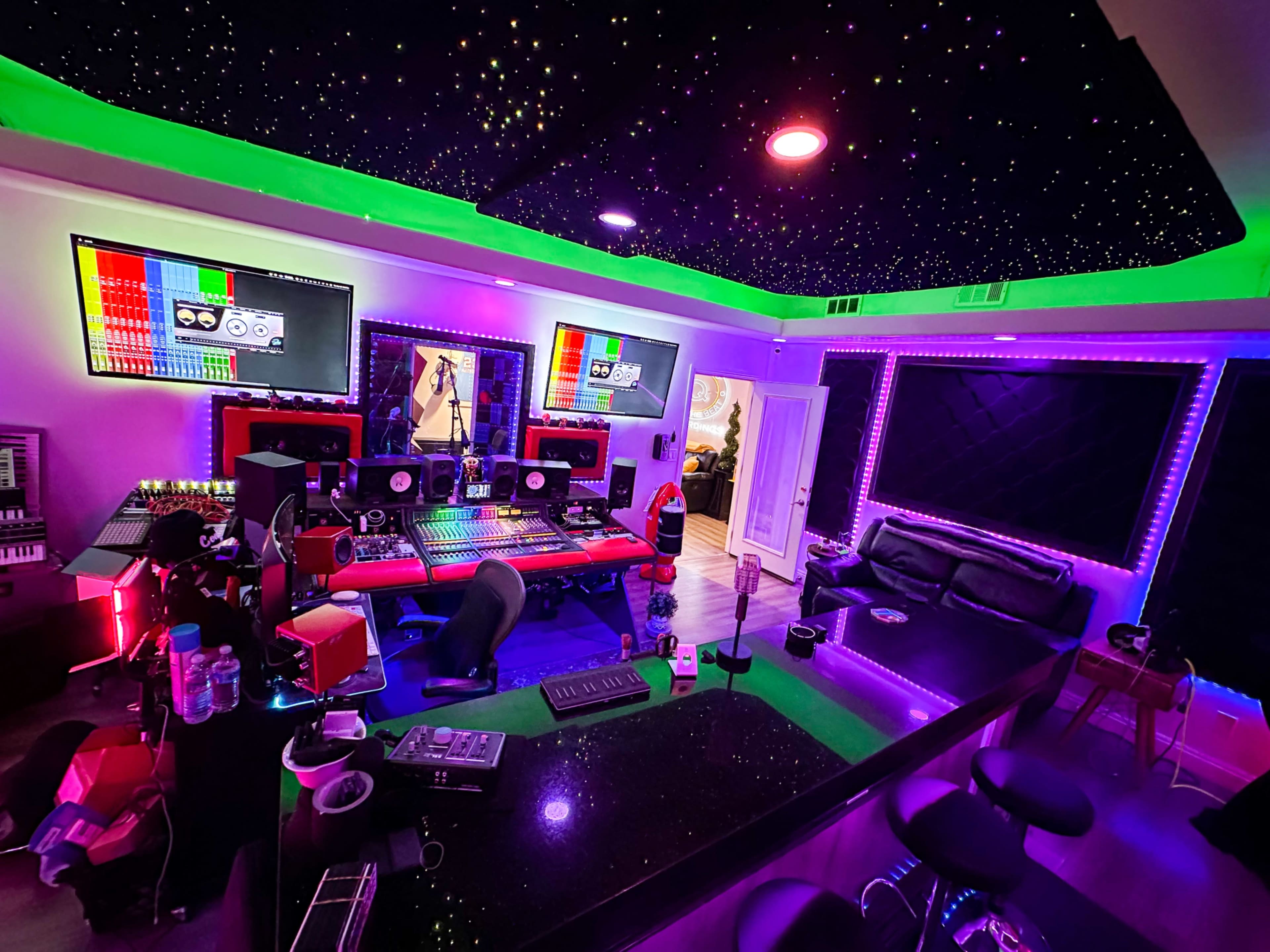 The image shows a music studio featuring a black countertop, multiple computer screens, red and green lighting, and a seating area with a black couch.