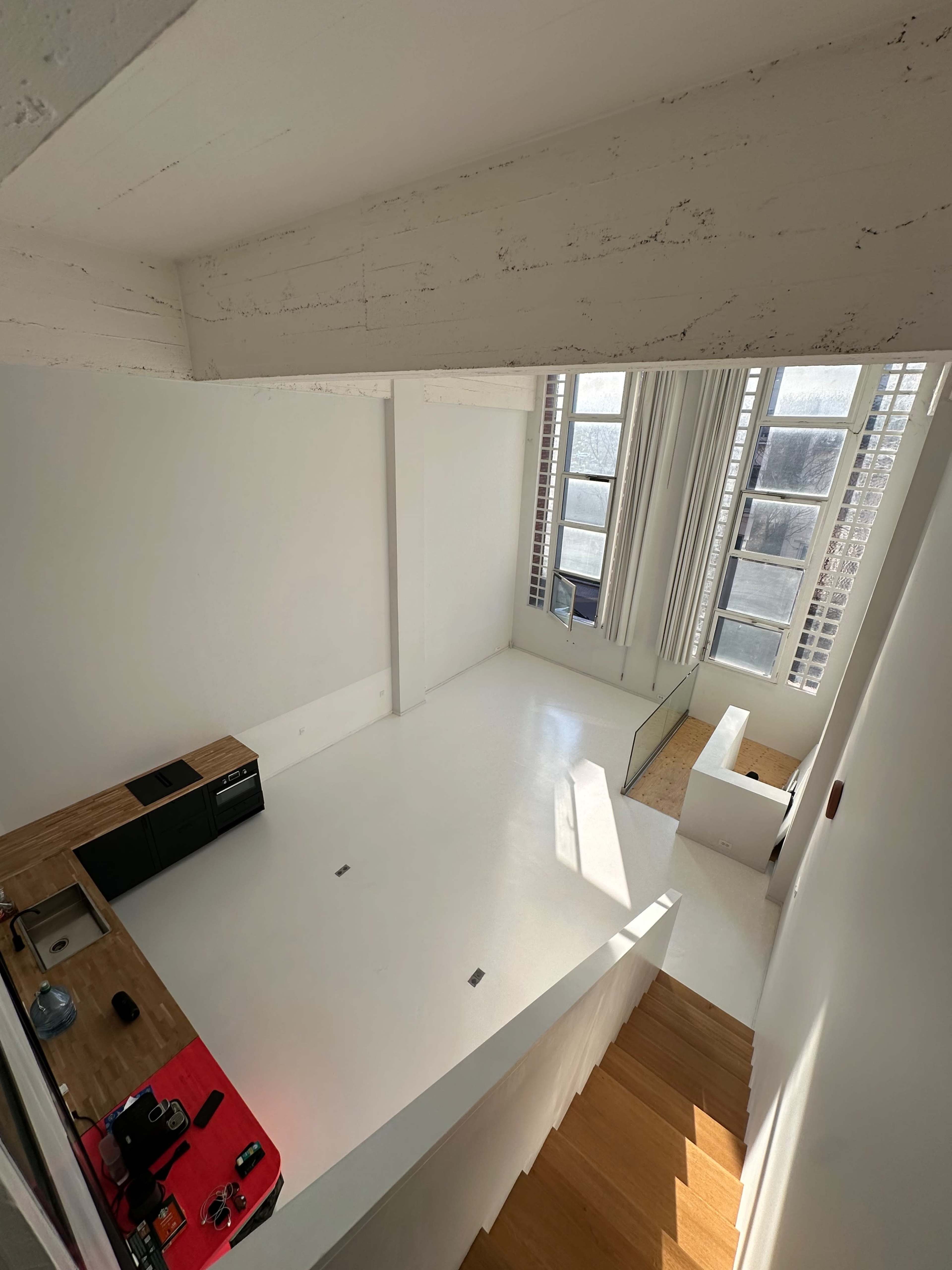 The image shows a spacious, minimalist interior with large windows, white walls, and a small kitchen area featuring a wooden countertop.