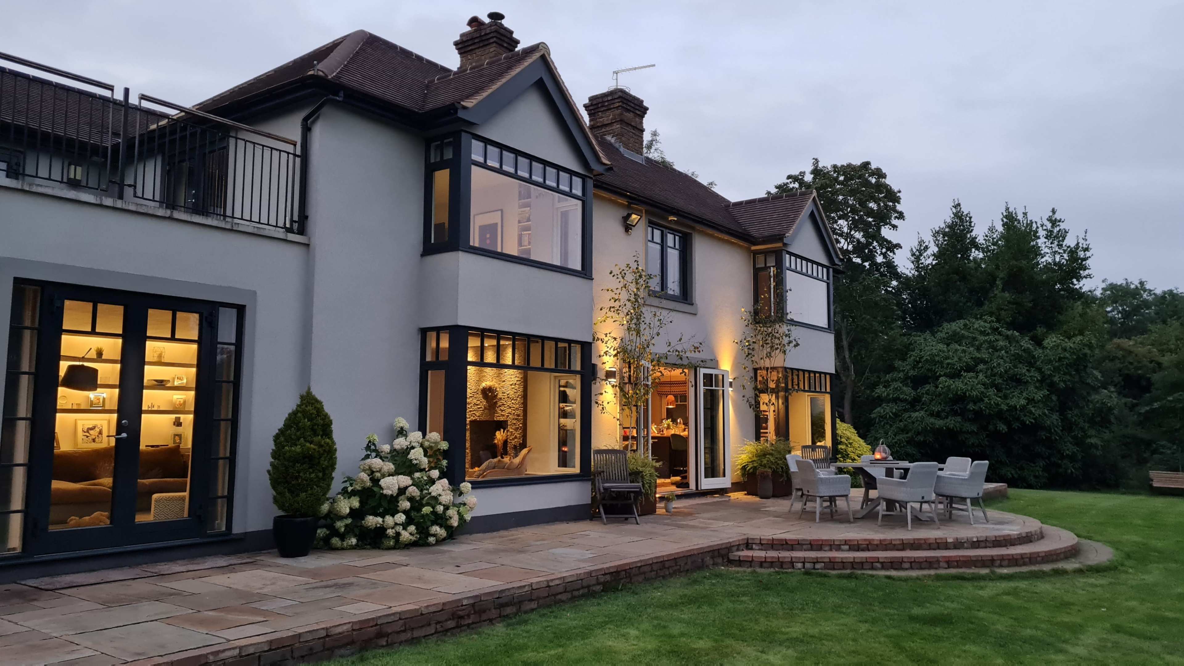 A two-story house with multiple large windows is set in a landscaped garden, featuring a patio with outdoor seating and illuminated interiors.