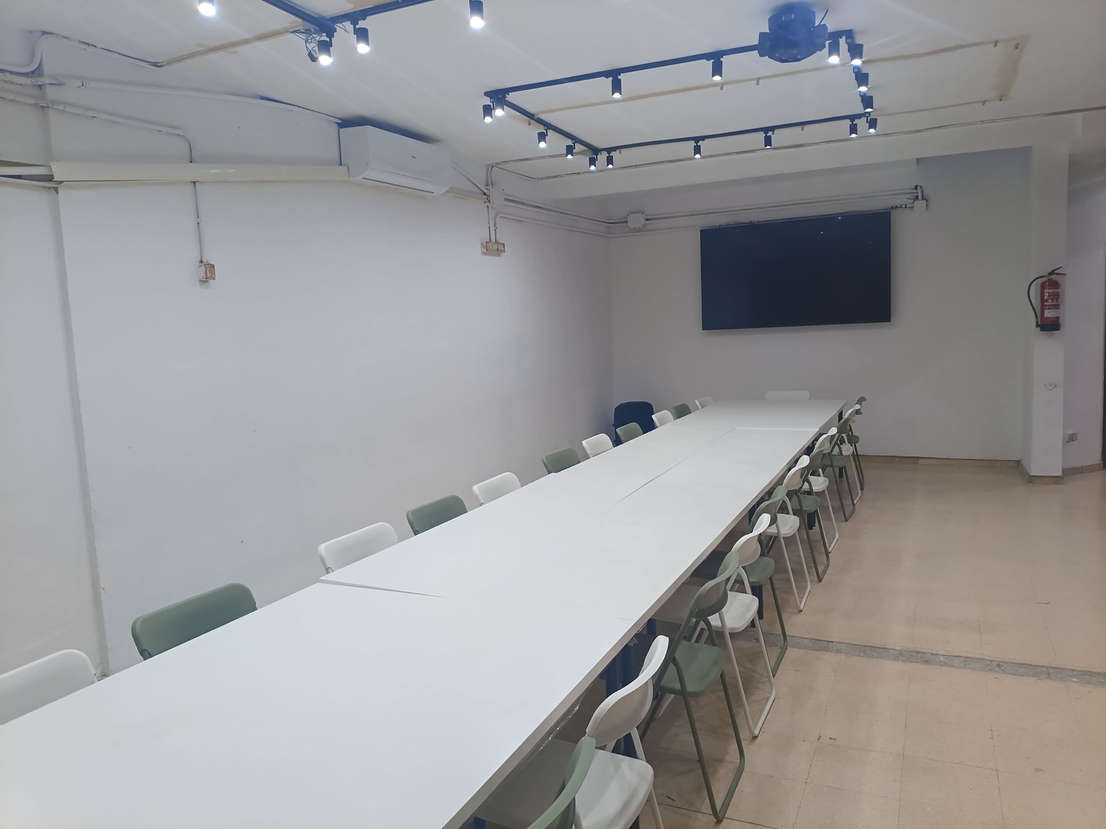 The image shows a long, rectangular conference table set in a brightly lit meeting room with chairs arranged on either side and a wall-mounted screen at one end.