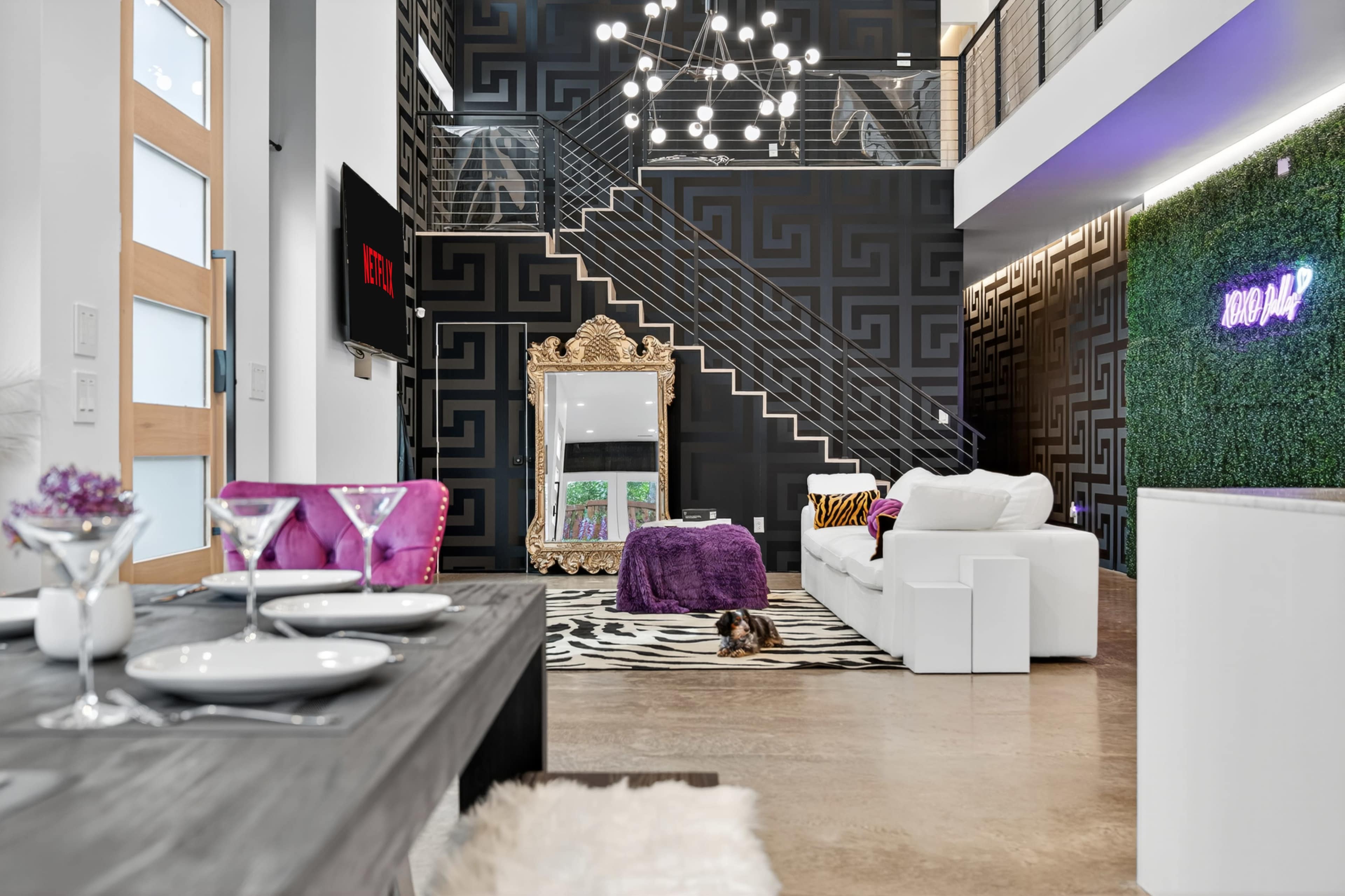 The image shows a modern living space with a staircase, a large mirror, a white couch, and decorative lighting on a patterned black wall.