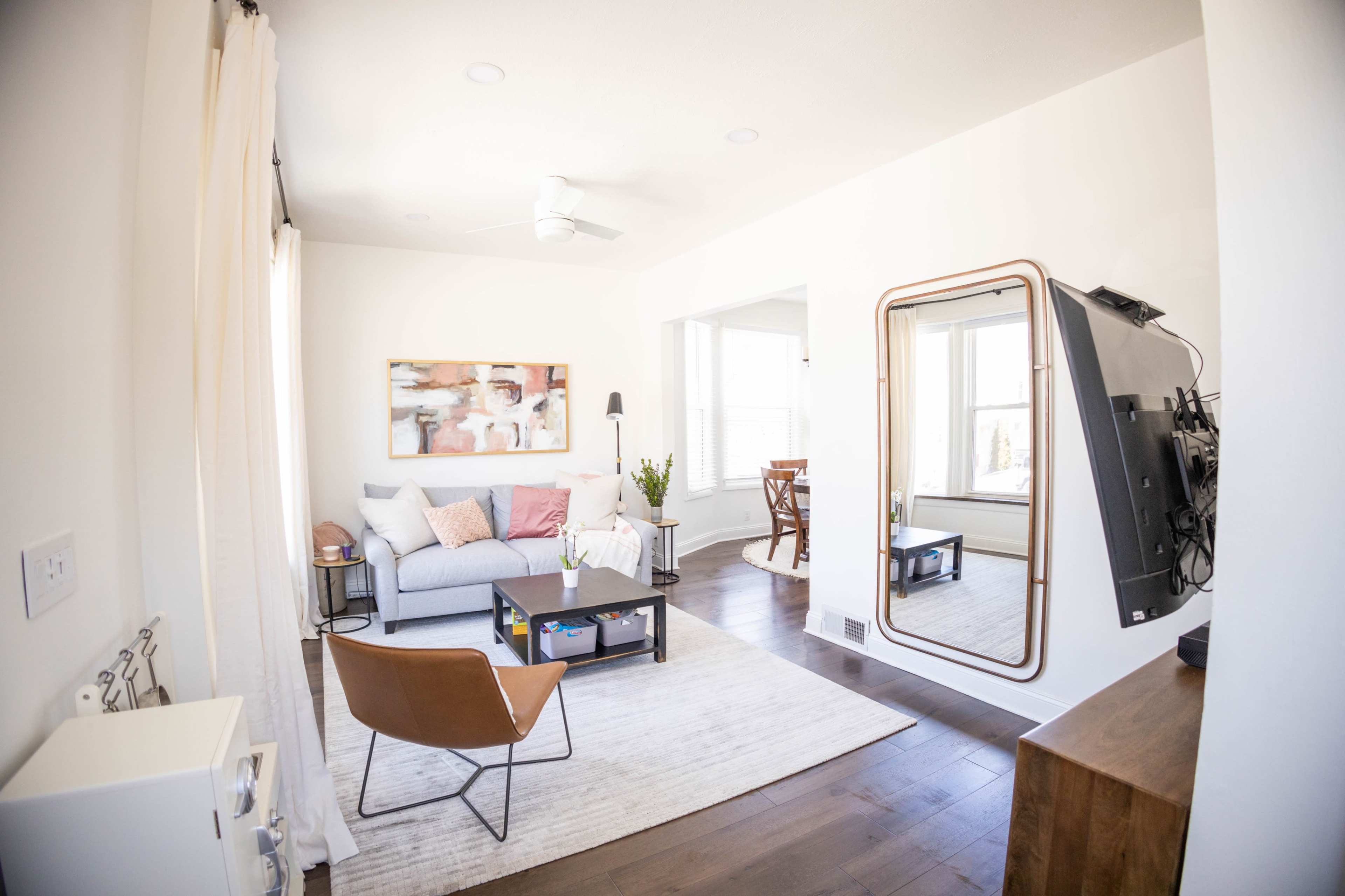 The image shows a bright living room with a gray sofa, a coffee table, a large mirror, and a mounted television.