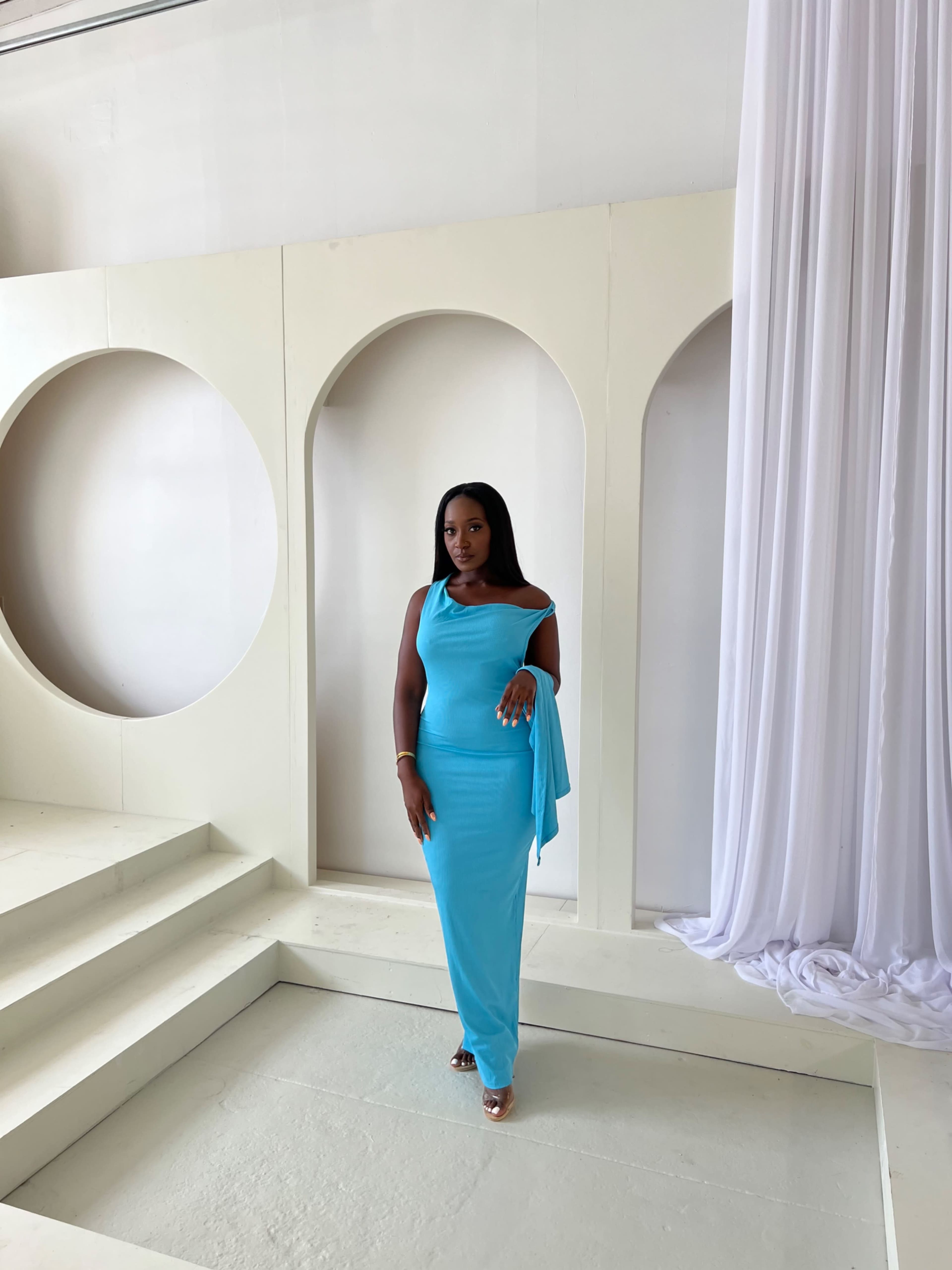 A woman stands elegantly on a set of white steps, wearing a form-fitting turquoise dress against a light-colored backdrop with arched cutouts.