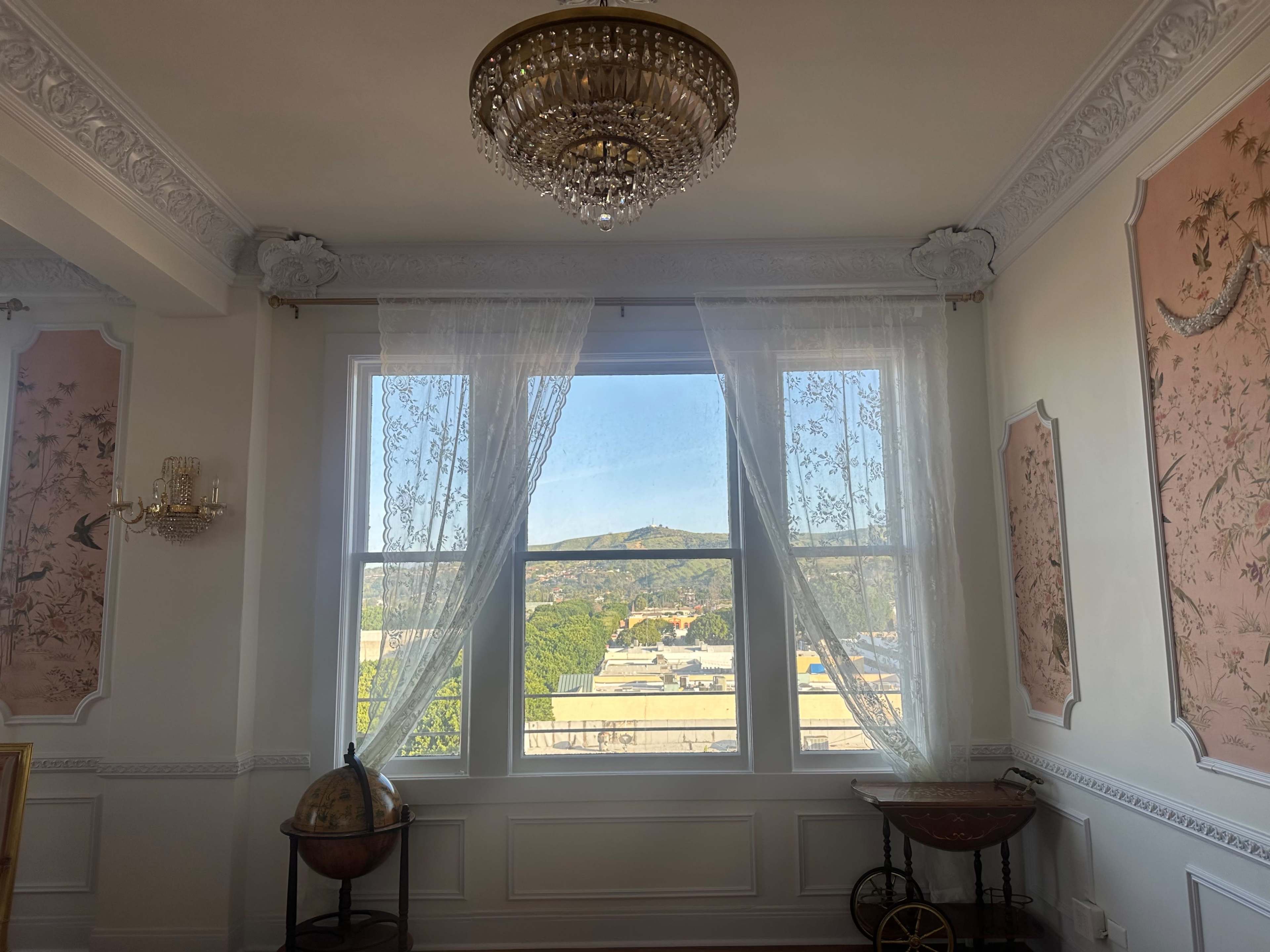 The image shows a well-lit room with large windows adorned with sheer curtains, framed by decorative wall art, and a view of greenery and hills outside.