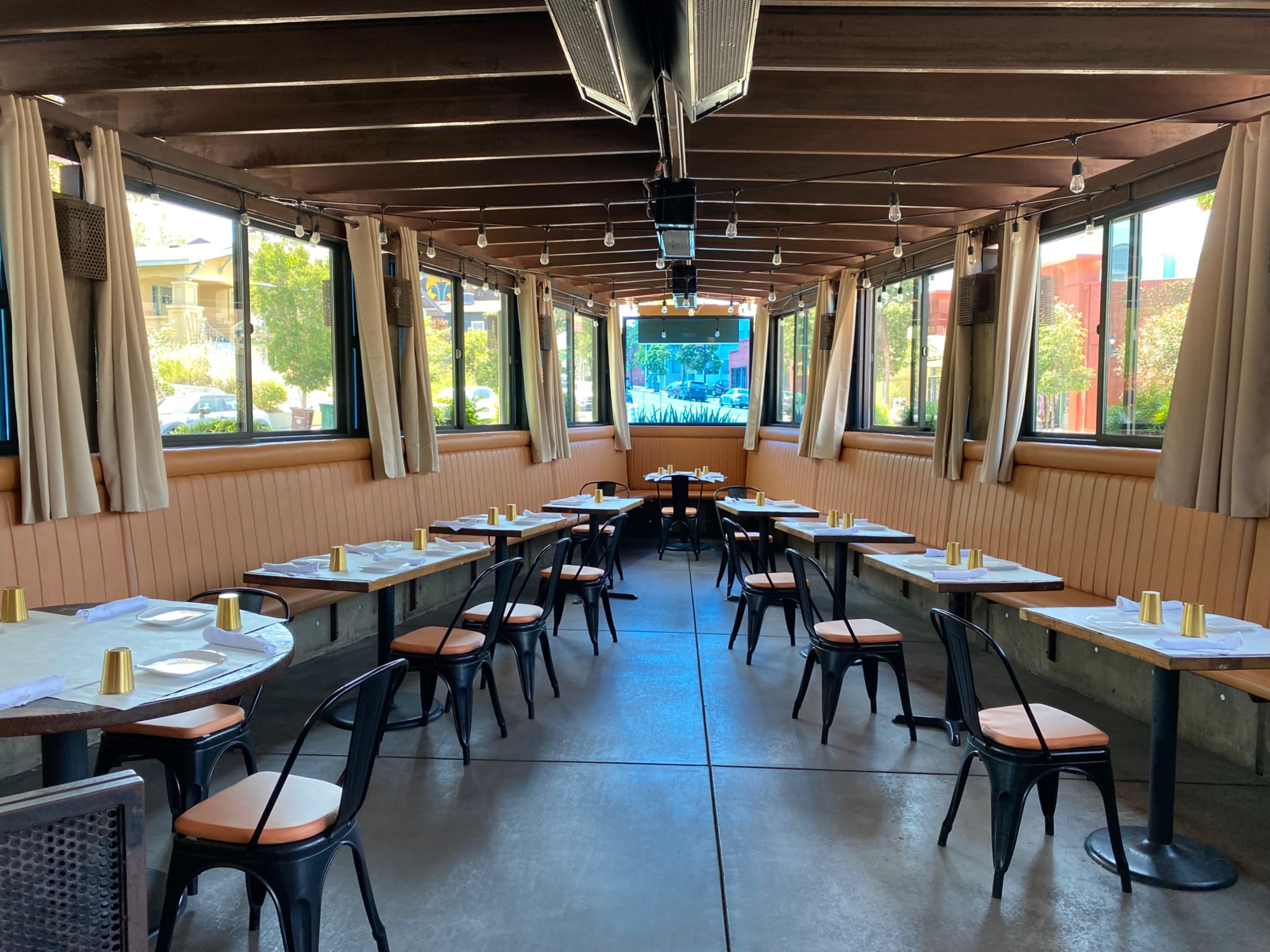 The indoor dining area features several tables with black metal chairs and gold accents, surrounded by large windows letting in natural light.