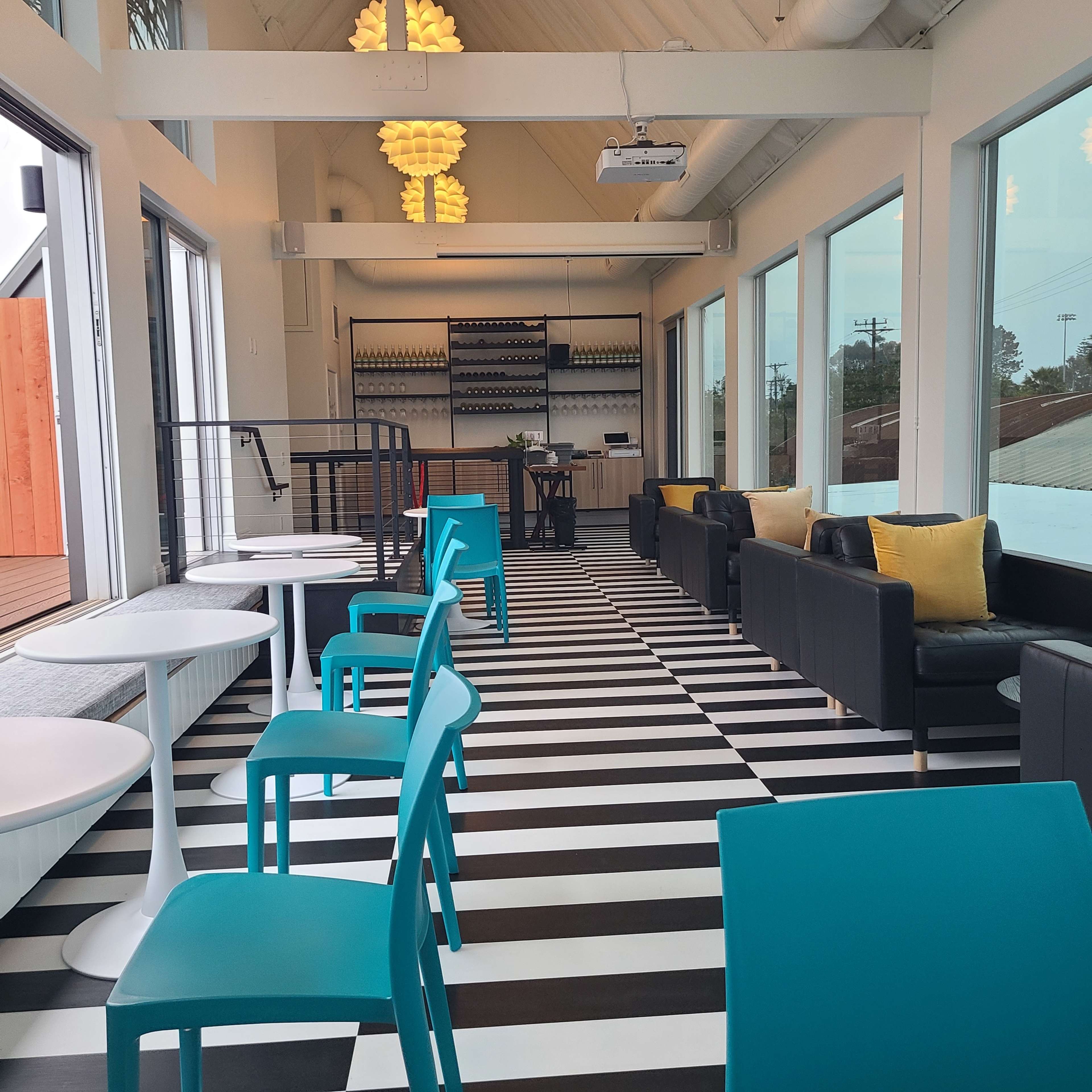 The image shows a modern indoor space with black and white striped flooring, featuring turquoise chairs, black couches with yellow pillows, and large windows allowing natural light.