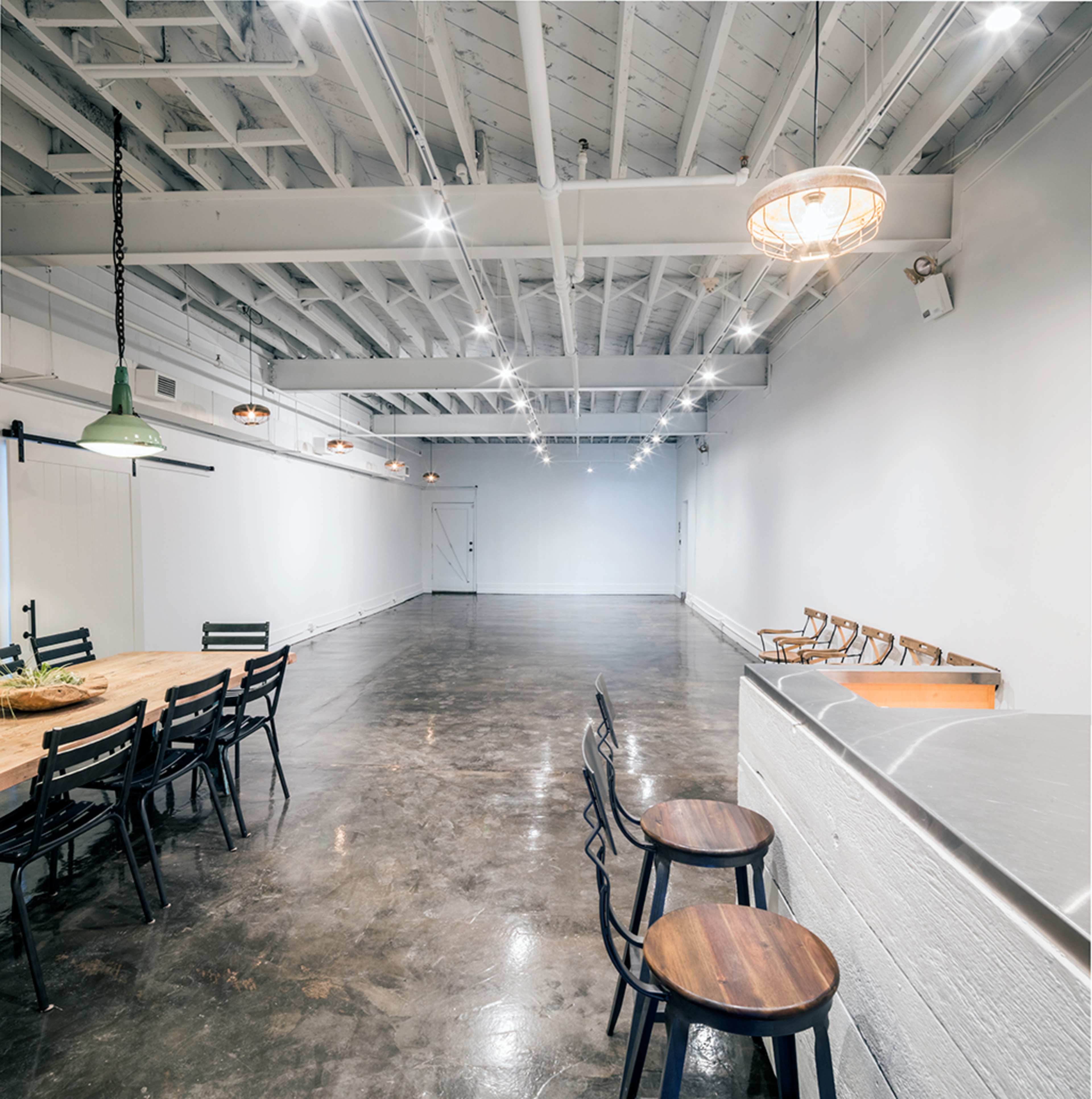 The image shows a spacious interior with polished concrete flooring, a long wooden table surrounded by black chairs, and a bar with wooden stools, all under a ceiling with exposed beams and pendant lighting.