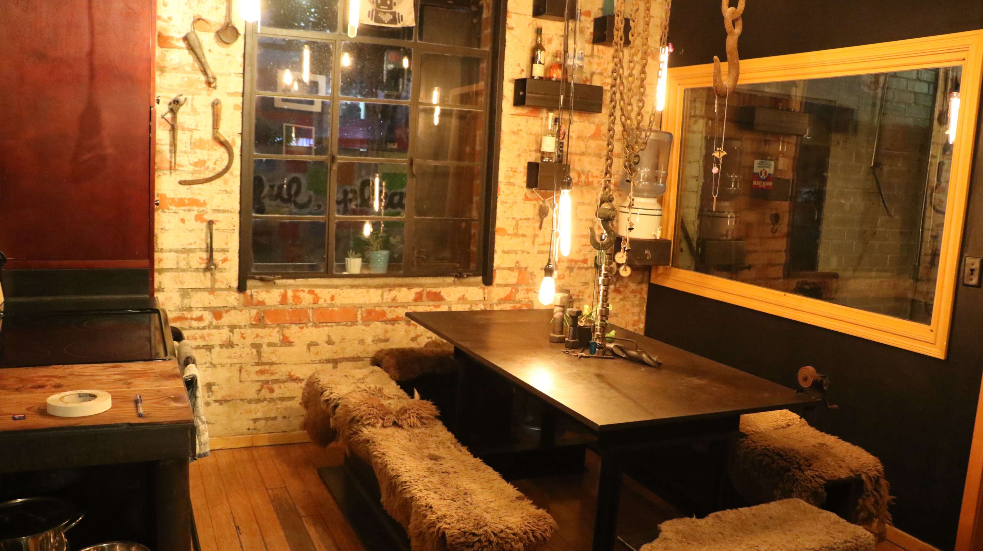 The image shows a rustic dining area with a wooden table surrounded by fur-covered benches, brick walls, and industrial lighting.