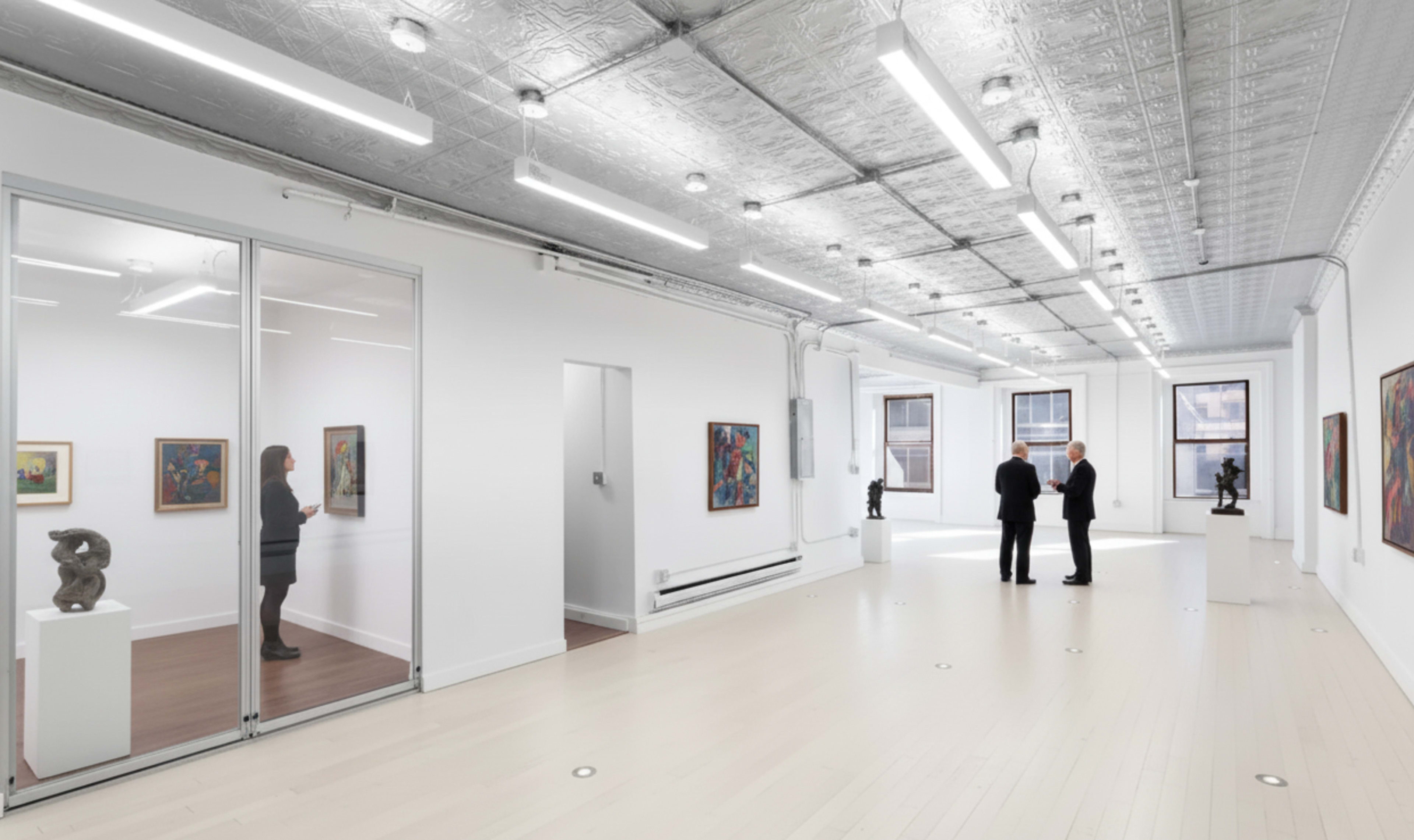 The image depicts a modern art gallery with two visitors conversing in the center while a woman stands in a nearby room observing artwork.