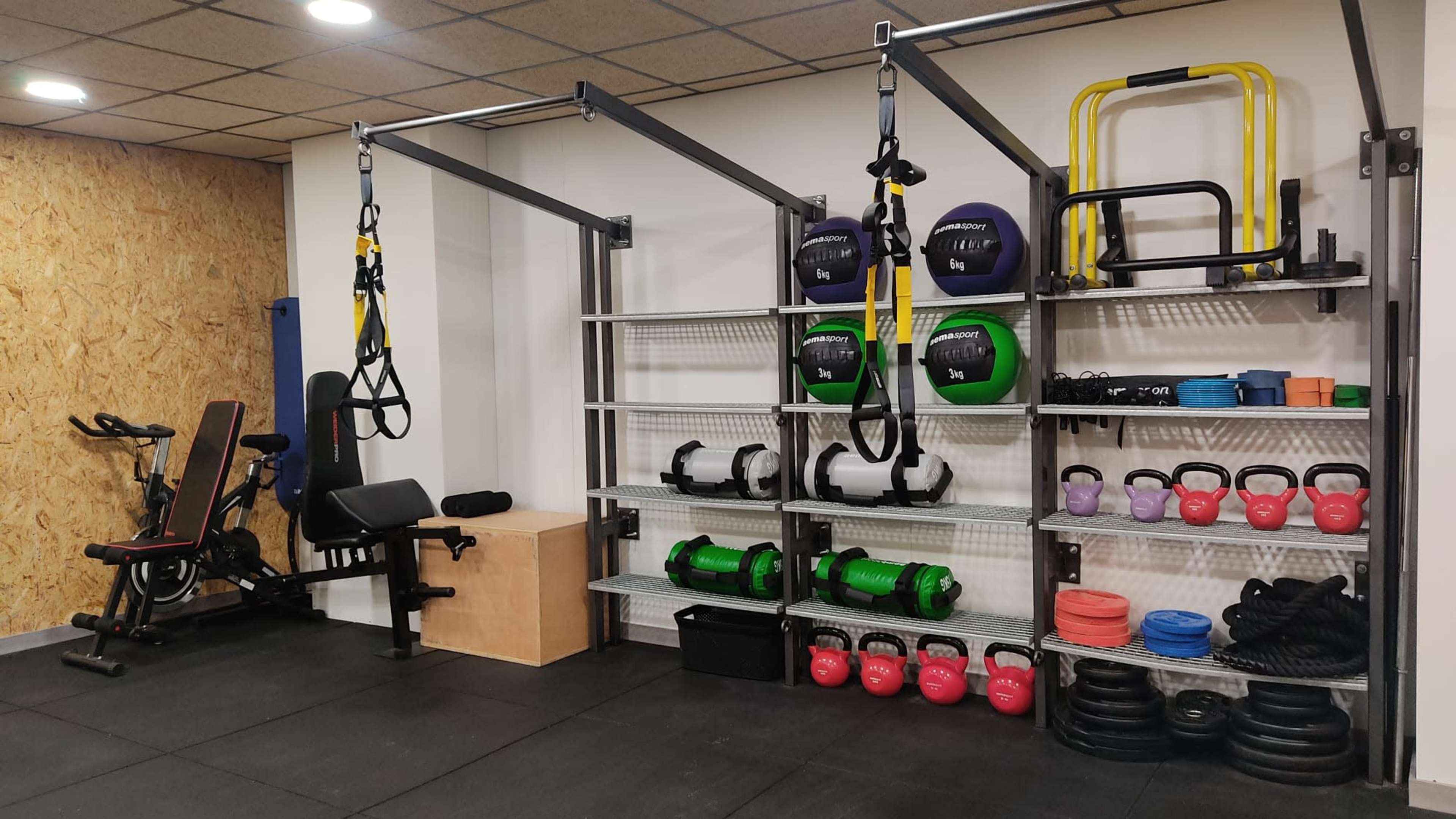 The image shows a gym setup featuring various exercise equipment, including weight plates, kettlebells, resistance bands, and a workout bench, organized on shelves and racks.