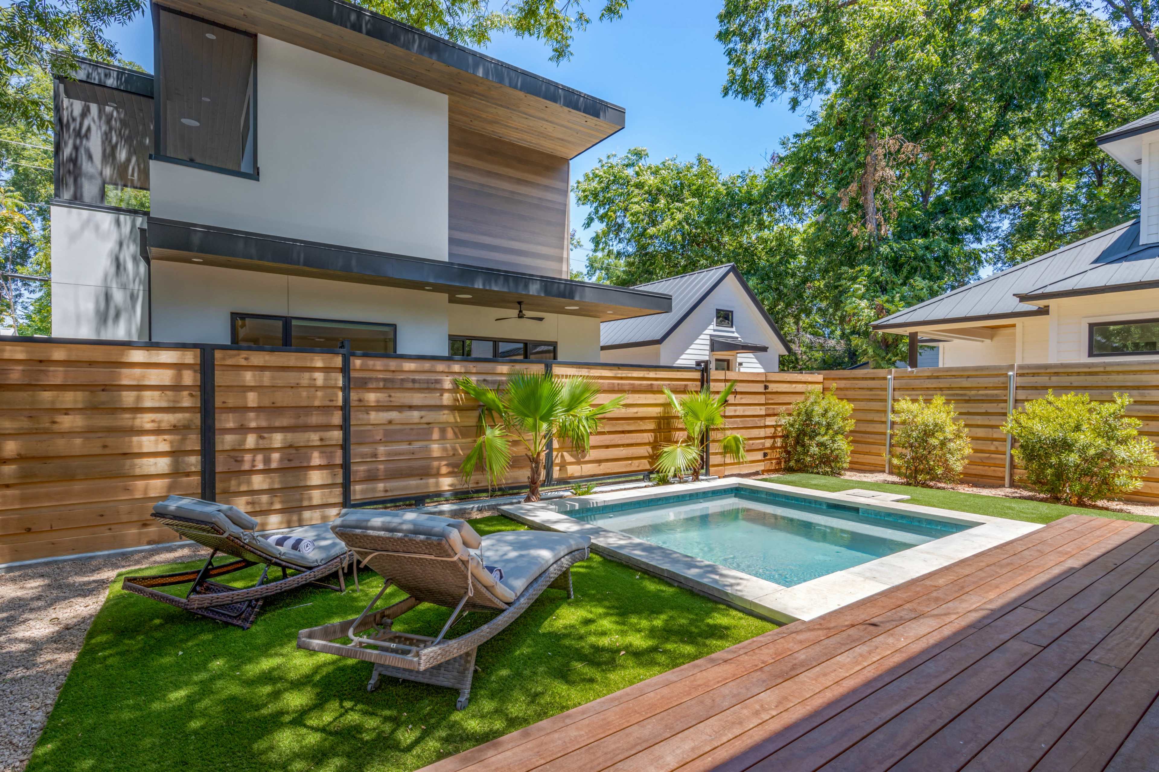 The image shows a modern backyard featuring a small pool surrounded by lounge chairs and landscaped greenery, with a wooden deck and a wooden fence enclosing the area.
