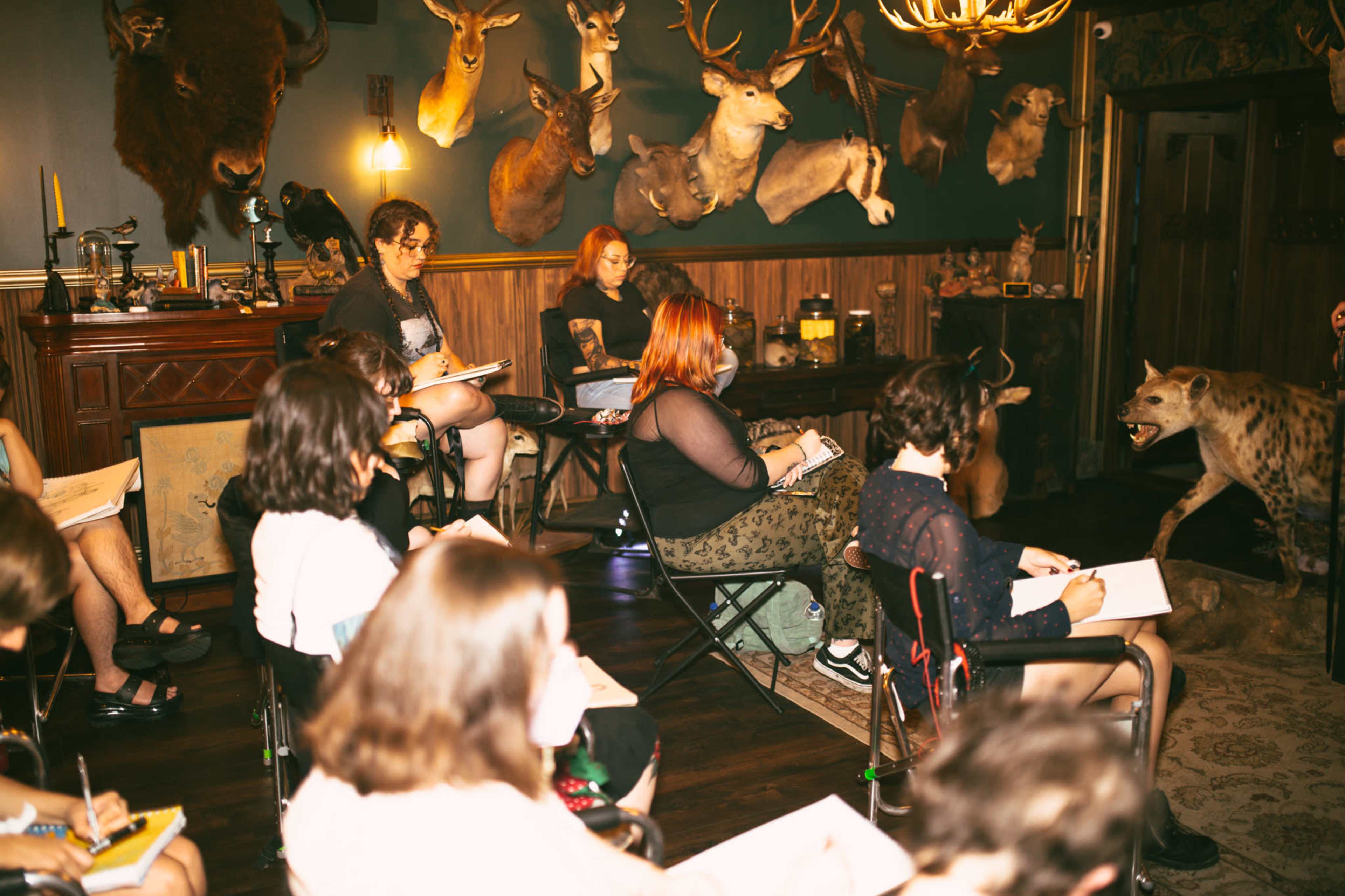 A group of people sits in a dark room filled with mounted animal heads, sketching while a hyena statue stands nearby.