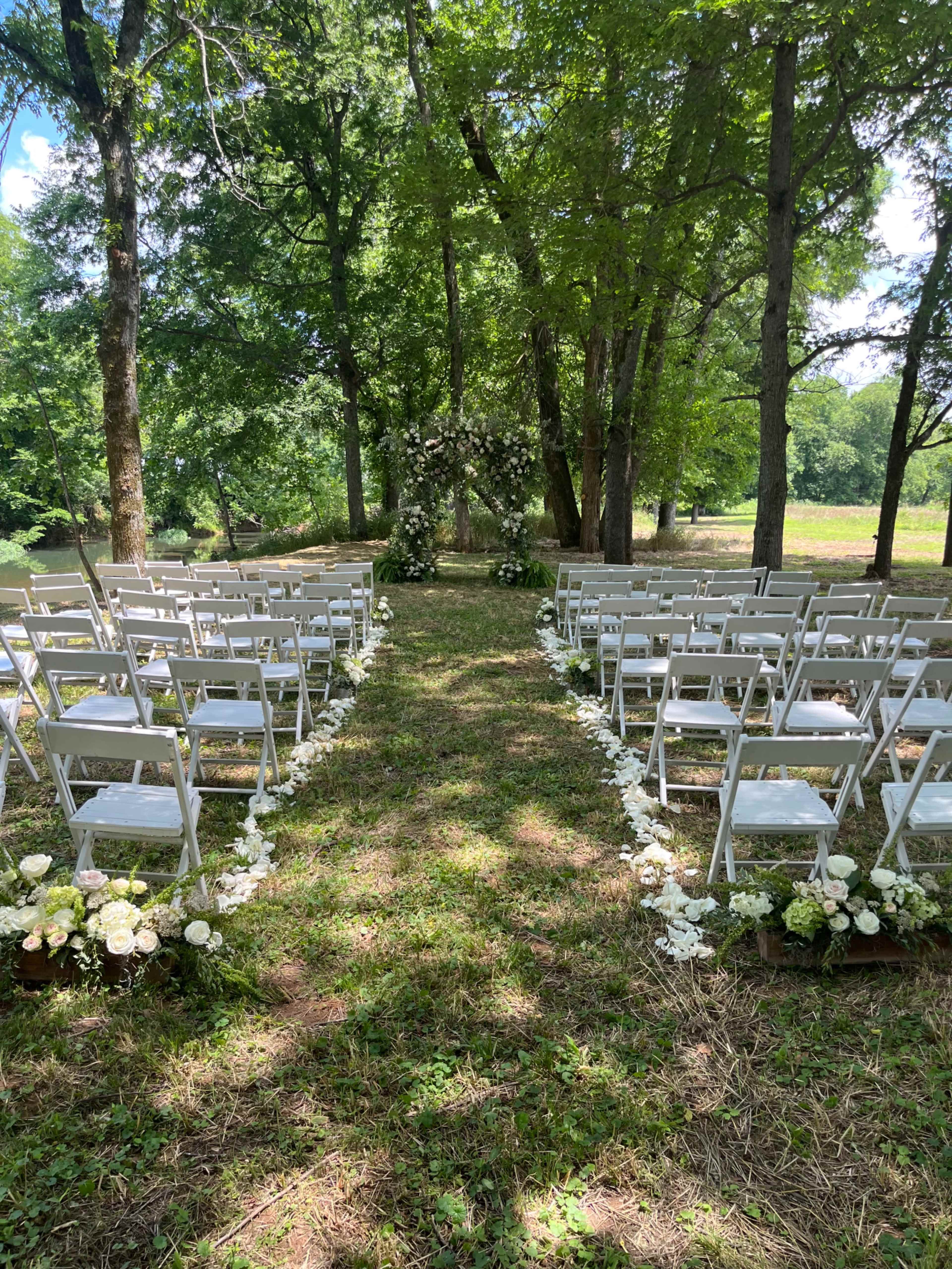 The image shows a wedding ceremony setup with white folding chairs arranged in two rows, leading to an arch adorned with flowers, all set in a wooded area.