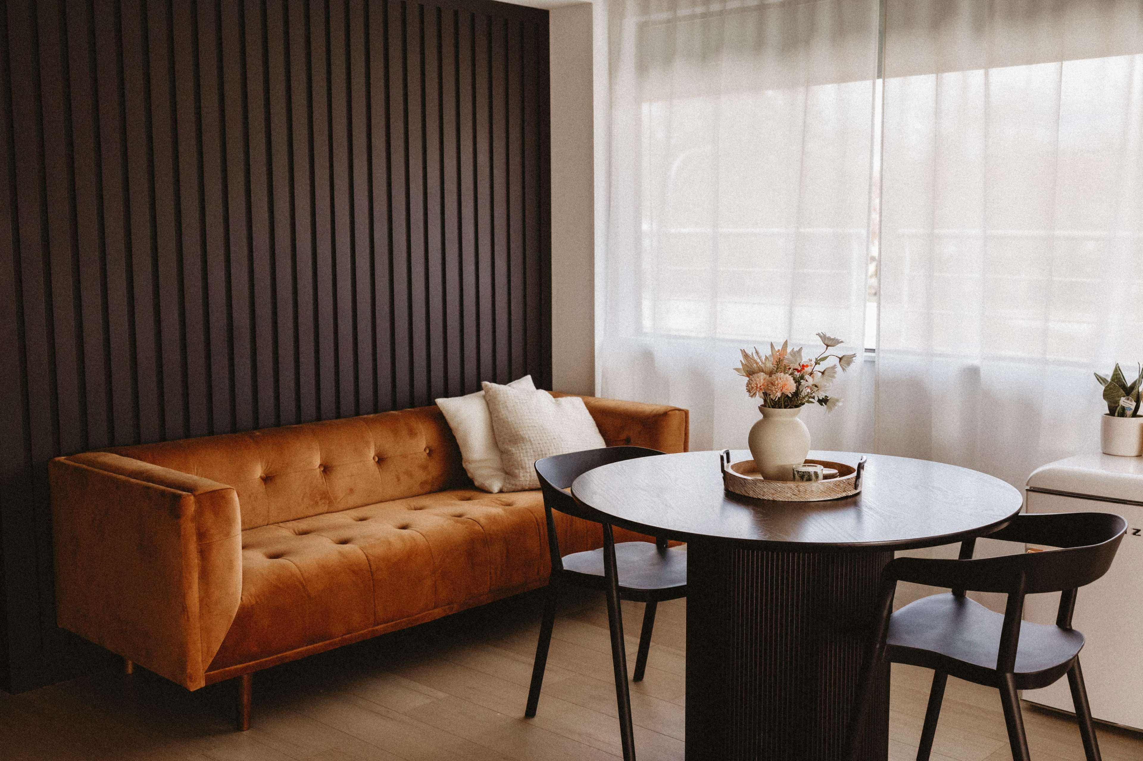 A modern dining area featuring a brown velvet sofa, a round black dining table, and a vase of flowers, with large windows allowing natural light to enter.