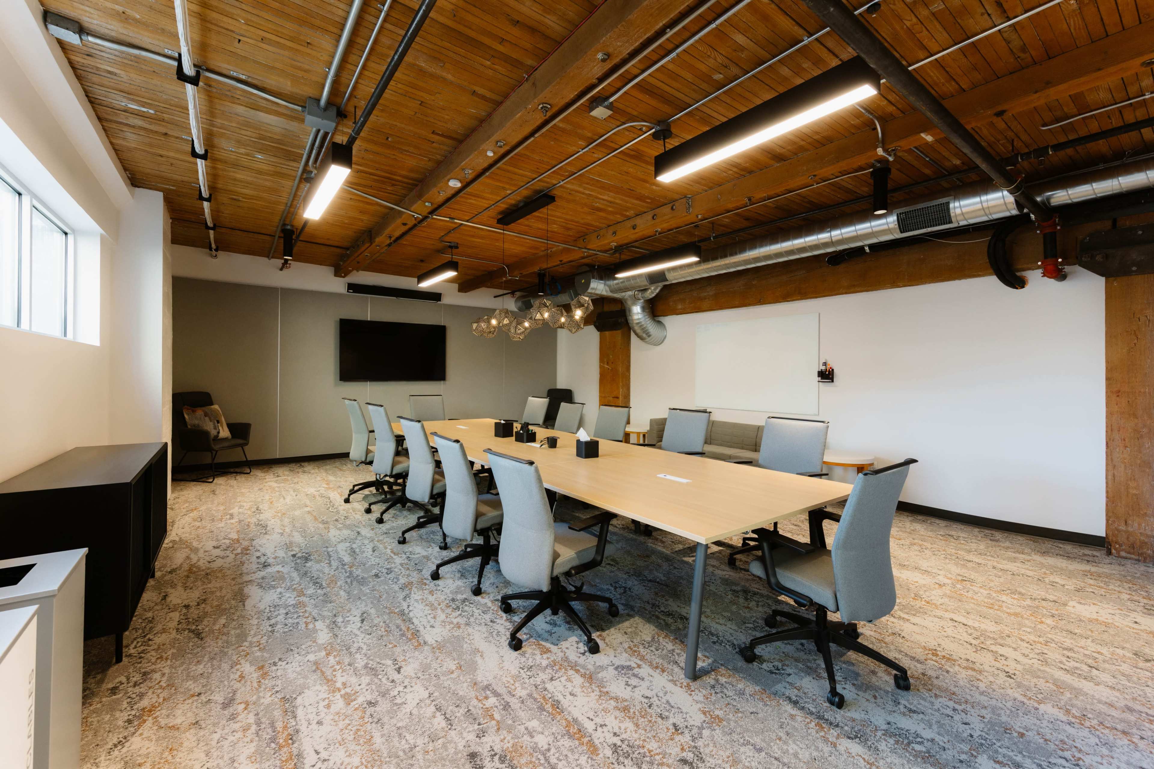 The image shows a modern conference room with a long wooden table, several ergonomic chairs, and a large screen on the wall.