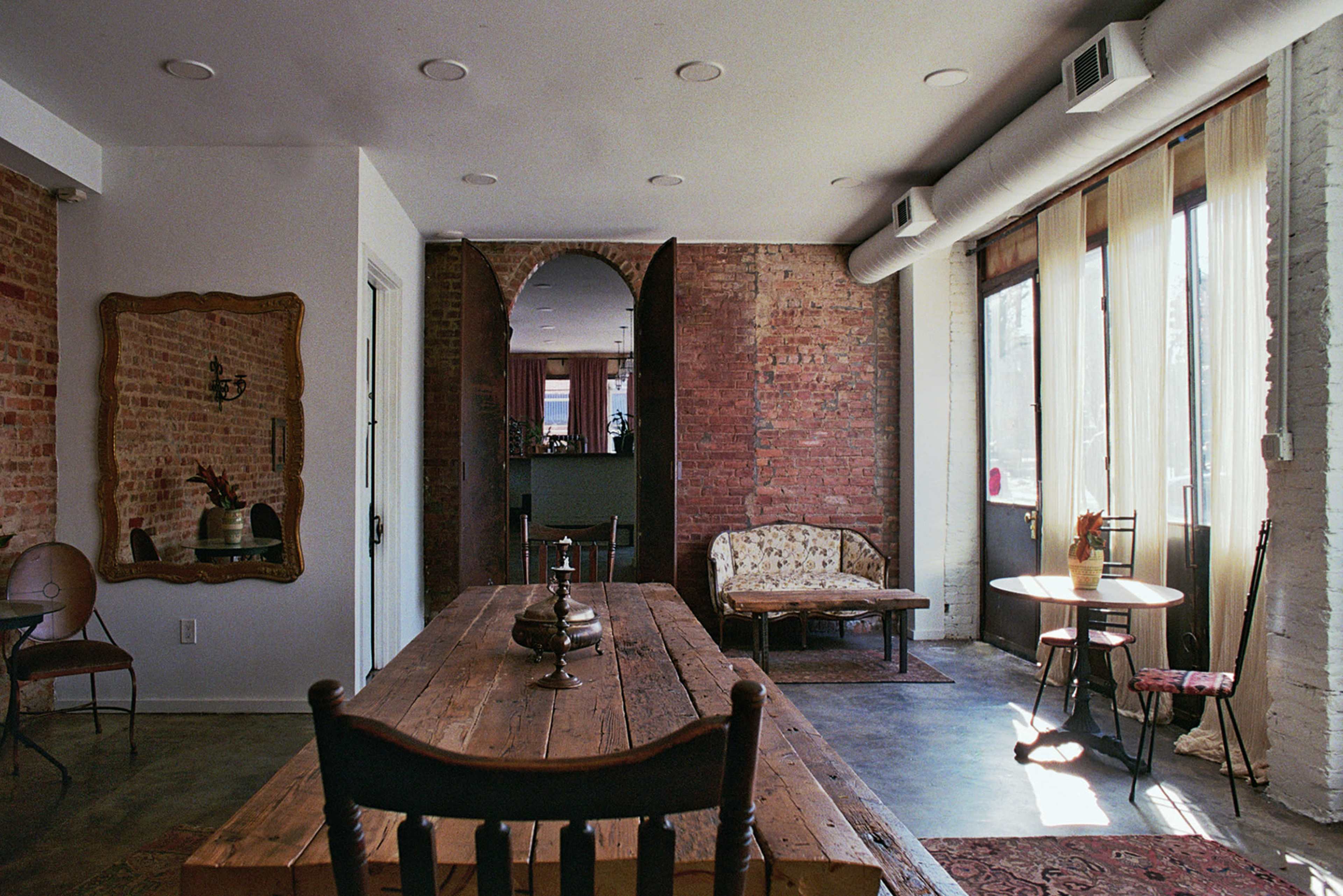 The image shows a stylish interior featuring exposed brick walls, a long wooden dining table, and a mix of seating options, including a vintage sofa and small round table, with large windows allowing natural light to enter the space.